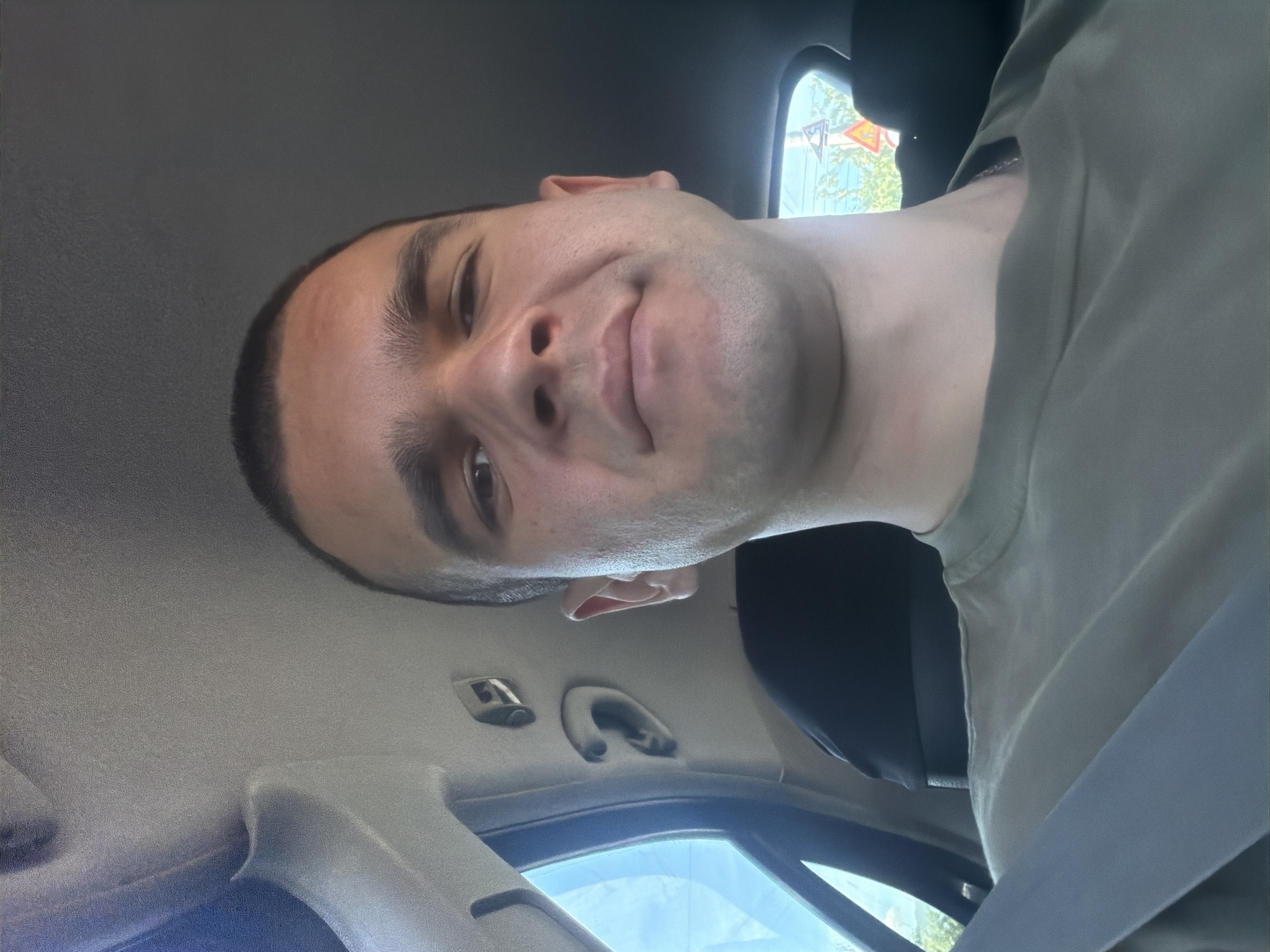 A man is in the driver's seat of a car. He has short dark hair, and is wearing a light gray T-shirt and a seatbelt. He appears to be taking a selfie. Through the car window, there are buildings and trees. Given the prompt about the location, it can be inferred that this image was taken somewhere within the city of Tulovo, Bulgaria. There are no specific details to identify a particular location within the city.