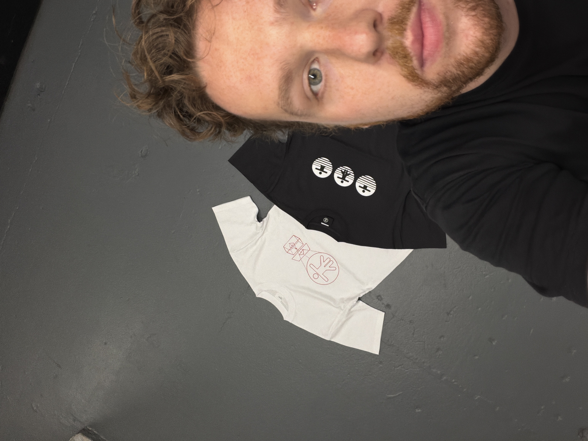 A person with fair skin, curly brown hair, blue eyes, and a beard is positioned in the upper right quadrant of the image. They are wearing a black t-shirt and are oriented so their face is prominent. To the left of the person are two t-shirts lying on a grey surface. The top t-shirt is black with a white graphic consisting of three circles with horizontal lines and an abstract design within each circle. Beneath this is a white t-shirt with a red abstract graphic, which appears to be an outline of a figure, a circle, and some geometric shapes. The grey surface has a slightly rough texture. The location and environment of the image cannot be definitively determined to be in New York City, United States of America without additional information.