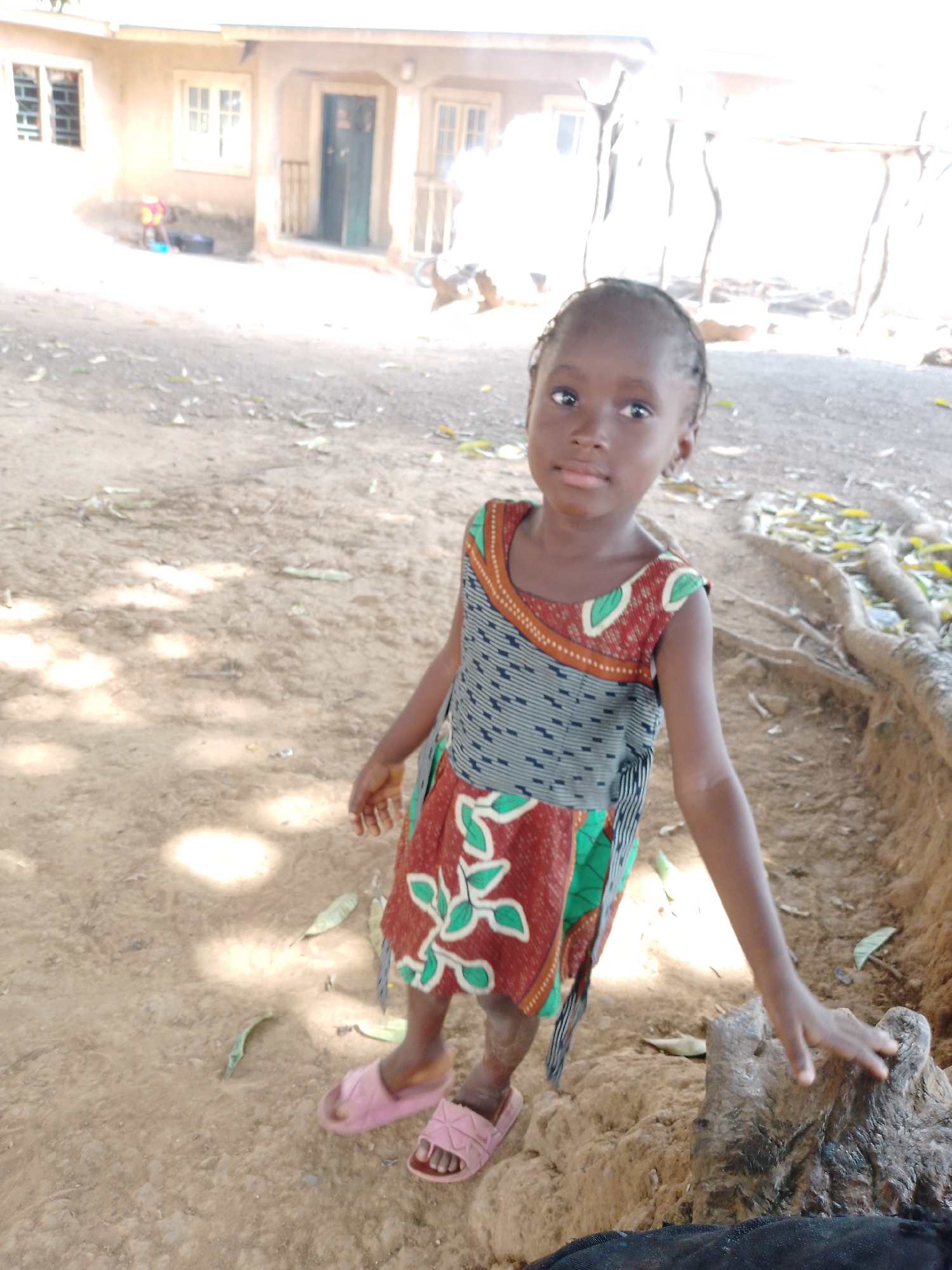 A young girl stands on a dirt surface outdoors. She wears a colorful sleeveless dress and pink sandals. The girl is positioned near a raised tree root and appears to be touching it. The background shows a building with windows and a door. The location is the city of Unknown, Unknown.