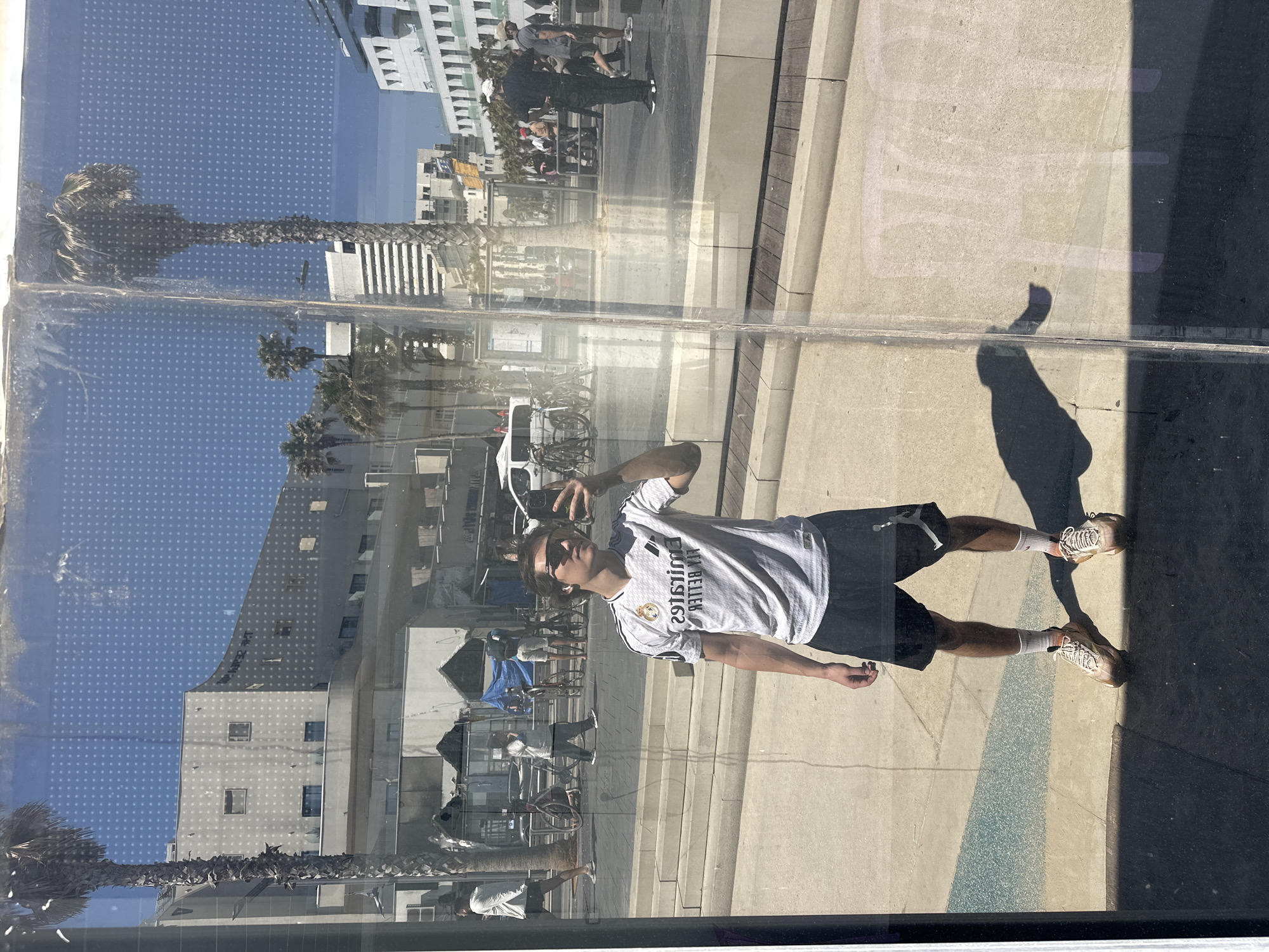 The image is a reflection of a person taking a selfie in a window, reflecting a street scene. The person is wearing a white Real Madrid soccer jersey, black shorts, and tan sneakers. They are holding a phone in their right hand.
The reflection shows a palm-lined street with buildings on the left, and people walking along the sidewalk. A white van is parked in the background. The sky is blue and sunny.
Based on the architectural style and the presence of palm trees, it is possible that the location is Tel Aviv, Israel. The details in the reflection such as architecture, trees, pavement and other elements would require more information to confirm this.