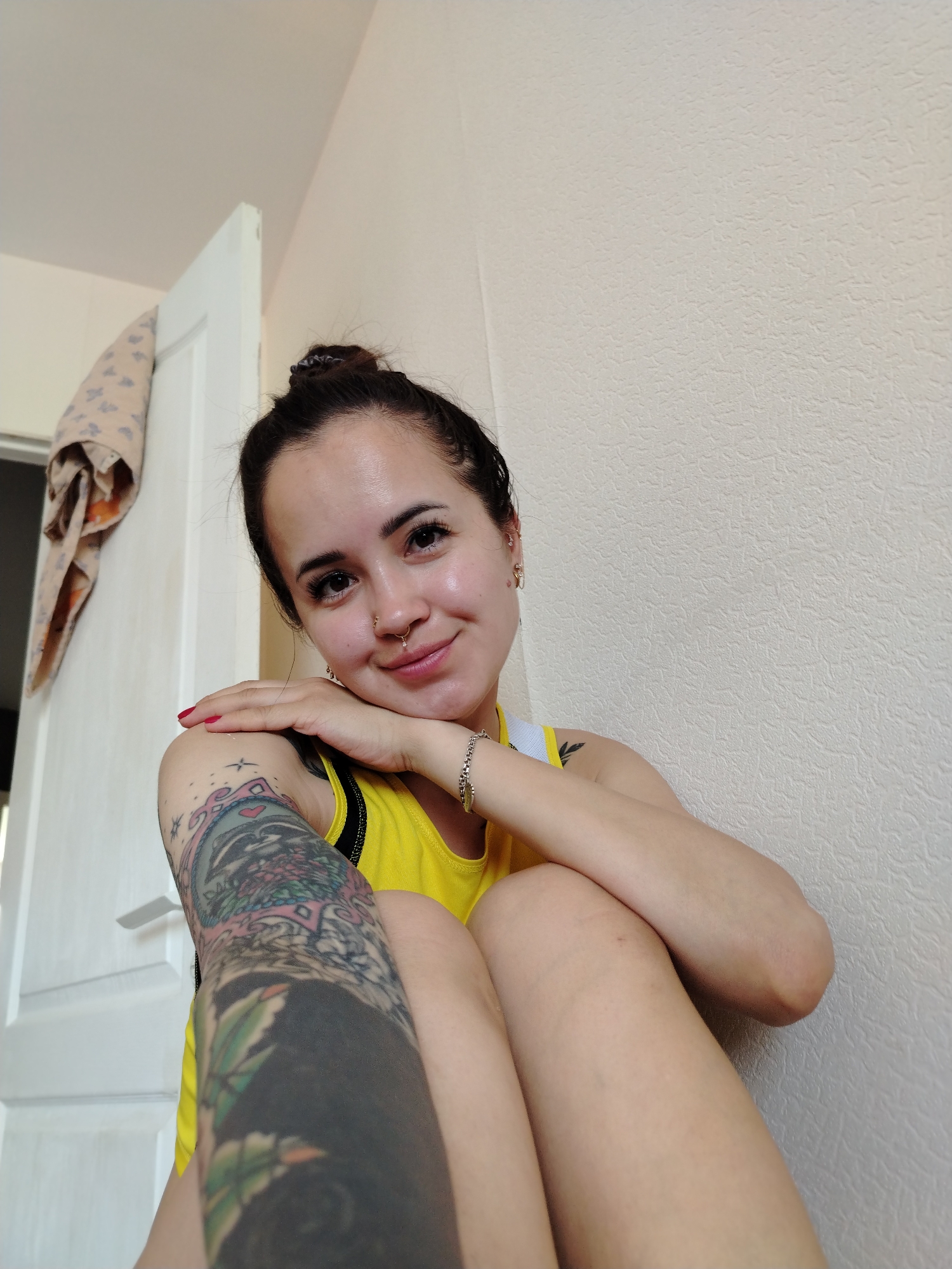 A young Caucasian woman with dark hair pulled into a bun and adorned with a nose ring and earrings, is seated indoors, leaning against a textured wall. She wears a yellow tank top and has extensive tattoos on her left arm, including a colorful design on her upper arm and a darker, more abstract pattern on her forearm. Her right arm is bent, with her hand resting on her shoulder. The background features a partially open white door with a patterned fabric hanging on it, and a plain, textured wall. The lighting is soft and natural. The location is identified as Noviy, Russia.
