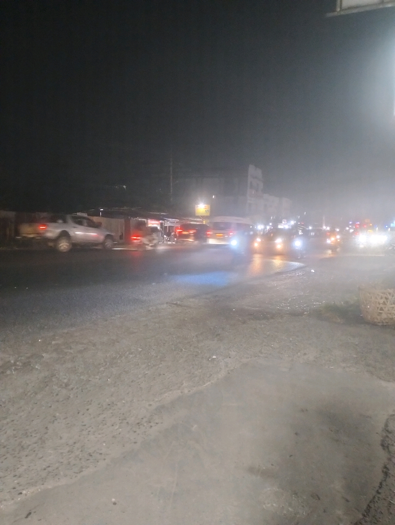 The image depicts a street scene at night, likely in Sebangar, Indonesia, characterized by a hazy or misty atmosphere that diffuses the light from vehicles and streetlights. The road surface appears wet, suggesting recent rain or high humidity.
Several vehicles are visible, primarily on the right side of the image. A silver pickup truck is prominent on the left, with its brake lights illuminated. Further down the road, a line of vehicles, including what appear to be vans or minibuses and other cars, are moving or stopped. Their headlights and taillights are bright and contribute to the diffused lighting effect. The vehicles suggest ongoing traffic flow.
Buildings or structures are visible in the background, illuminated by streetlights and vehicle lights, but their details are obscured by the atmospheric conditions. There are no clearly discernible individuals in the scene, though the presence of vehicles implies human activity.
On the far right of the image, a woven basket or similar object is partially visible, suggesting a possible market or roadside stall setup. The overall impression is that of a bustling but somewhat obscured nighttime urban environment.