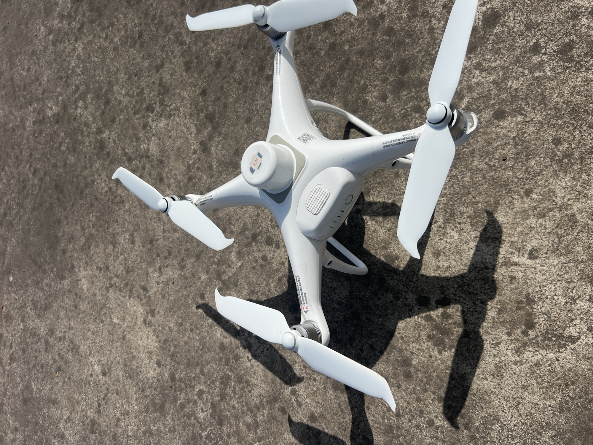 A white drone is laying on a concrete surface, likely outdoors. The drone has four propellers, each with two blades. There is Chinese writing on the drone near the base of the propellers. There is a small cylindrical component on top of the drone with the letters "I/IIM" printed on it. A shadow of the drone is visible on the concrete surface. The concrete appears worn and uneven. There are no people visible in the image. The scene is not definitively located in Jiangjin, China; however, the Chinese writing on the drone implies that the item is either manufactured or intended for use in China.