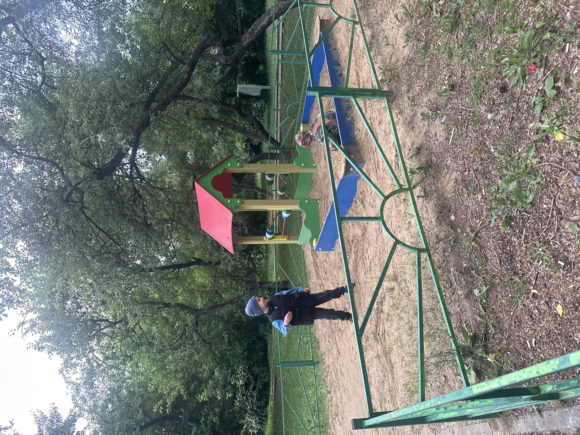In Lytkarino, Russia, a children's playground is depicted on a cloudy day, surrounded by lush green trees and a light-colored fence. Two young children are present: one boy, wearing a blue hat, black vest, blue shirt, and dark pants, stands near the foreground. The second child, wearing a camouflage-patterned shirt and hat, is seated in a sandpit. A colorful play structure, featuring a red roof and yellow supports with blue and green accents, is situated in the sandpit. The ground is covered with sand and scattered debris.