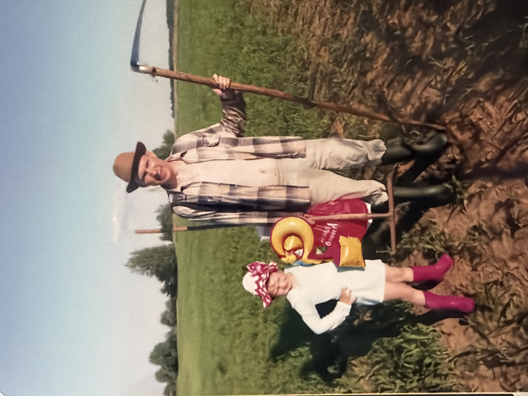 The image depicts a man and a child standing in a field. The man is wearing a hat, a plaid shirt, and tall rubber boots, and he is holding a scythe over his shoulder. The child is wearing a white dress, pink boots, and a headscarf with a red and white bow. There is a red bag and a yellow inflatable ring sitting on a seat near the child. The field is green, and there is a clear blue sky. There is no indication that this photograph was taken in a city; the background is agricultural. The specific location is unknown.