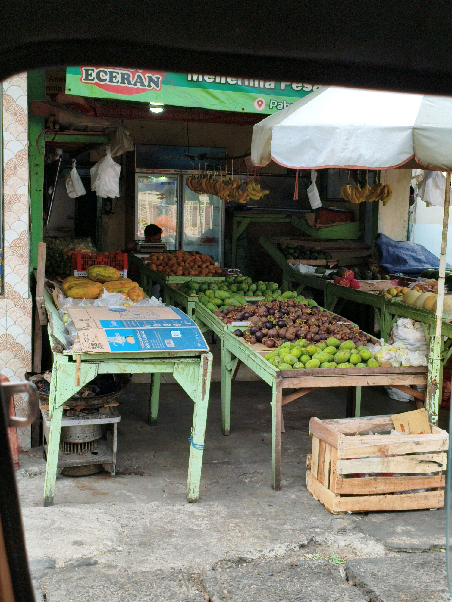 The image depicts an outdoor fruit stand in Cibinong, Indonesia. The stall features several wooden tables painted light green, displaying a variety of fruits. In the foreground, a light green table holds papayas and what appear to be large yellow gourds. A cardboard box with blue and white printing sits on this table. To the right, a wooden crate filled with plastic bags is visible on the ground. Behind these, other tables are laden with green mangoes, purple mangosteens, and piles of smaller, round orange fruits. Bananas are hanging from the stall's awning in several bunches. The background shows the interior of the shop with more fruit displays and a person visible behind a counter. An umbrella provides shade over part of the stall. The ground is a concrete pavement with some cracks and debris. The overall scene is that of a bustling local market.