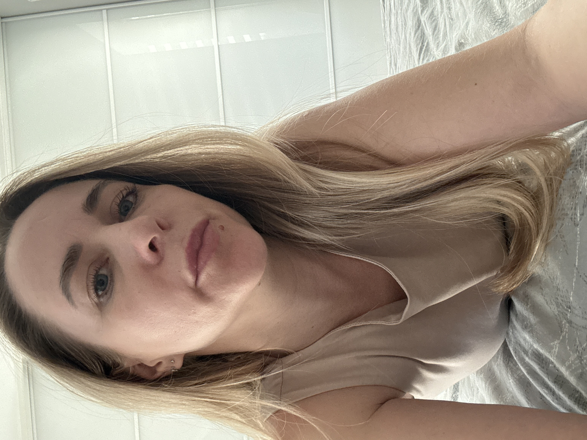 A young woman with long, wavy blonde hair and blue eyes is the central subject of this sideways-oriented selfie. She is depicted lying down, likely on a bed or sofa, indicated by the grey and white patterned fabric resembling a duvet or blanket on the right side of the image. She is wearing a simple, light beige or nude-colored sleeveless top. Her gaze is directed at the camera with a neutral and relaxed expression.
Notable details include a small mole above her upper lip on the right side of her face, and two delicate silver-colored earrings in her visible left ear. Her fair skin shows fine arm hair and a small, dark mole on her forearm. The background features a light-colored, possibly white, wall with subtle vertical panel lines, suggesting an indoor setting such as a bedroom or living area. The lighting is soft and natural, implying daytime. No visible text is present. The context provided indicates the location is Chaikovsky, Russia.