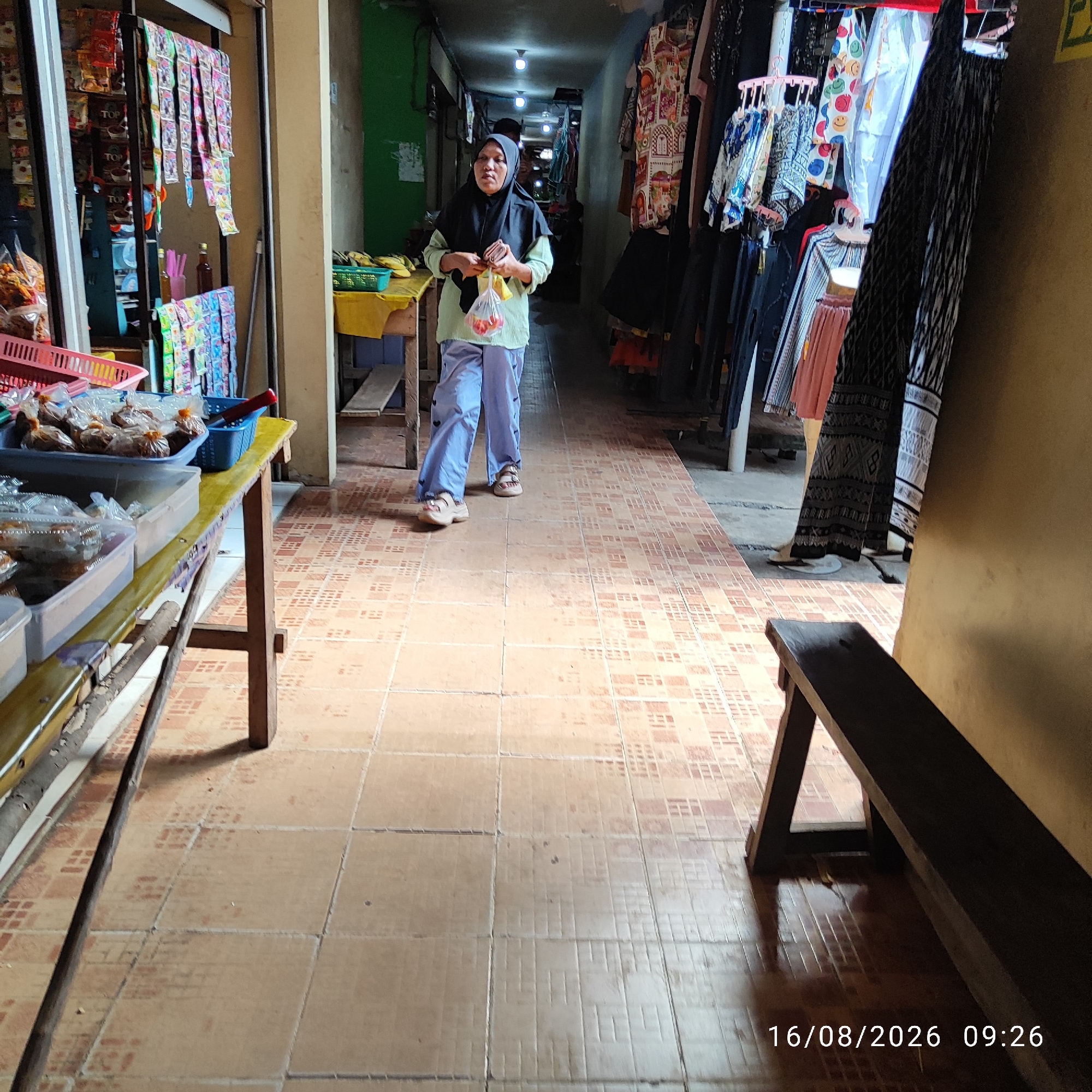 The image depicts a narrow marketplace aisle in what appears to be Pembaharuan, Indonesia. The aisle is lined with stalls selling various goods. On the left, a stall displays packaged snacks and drinks. Further down the aisle on the right, clothing is hung for sale. A woman wearing a headscarf and a light-colored blouse with light blue pants is walking away from the camera towards the deeper end of the aisle. She is holding a plastic bag containing red fruits, possibly tomatoes. The floor is tiled with a patterned terracotta tile. A wooden bench is visible in the foreground on the right side of the aisle. The timestamp "16/08/2026 09:26" is visible in the bottom right corner of the image.