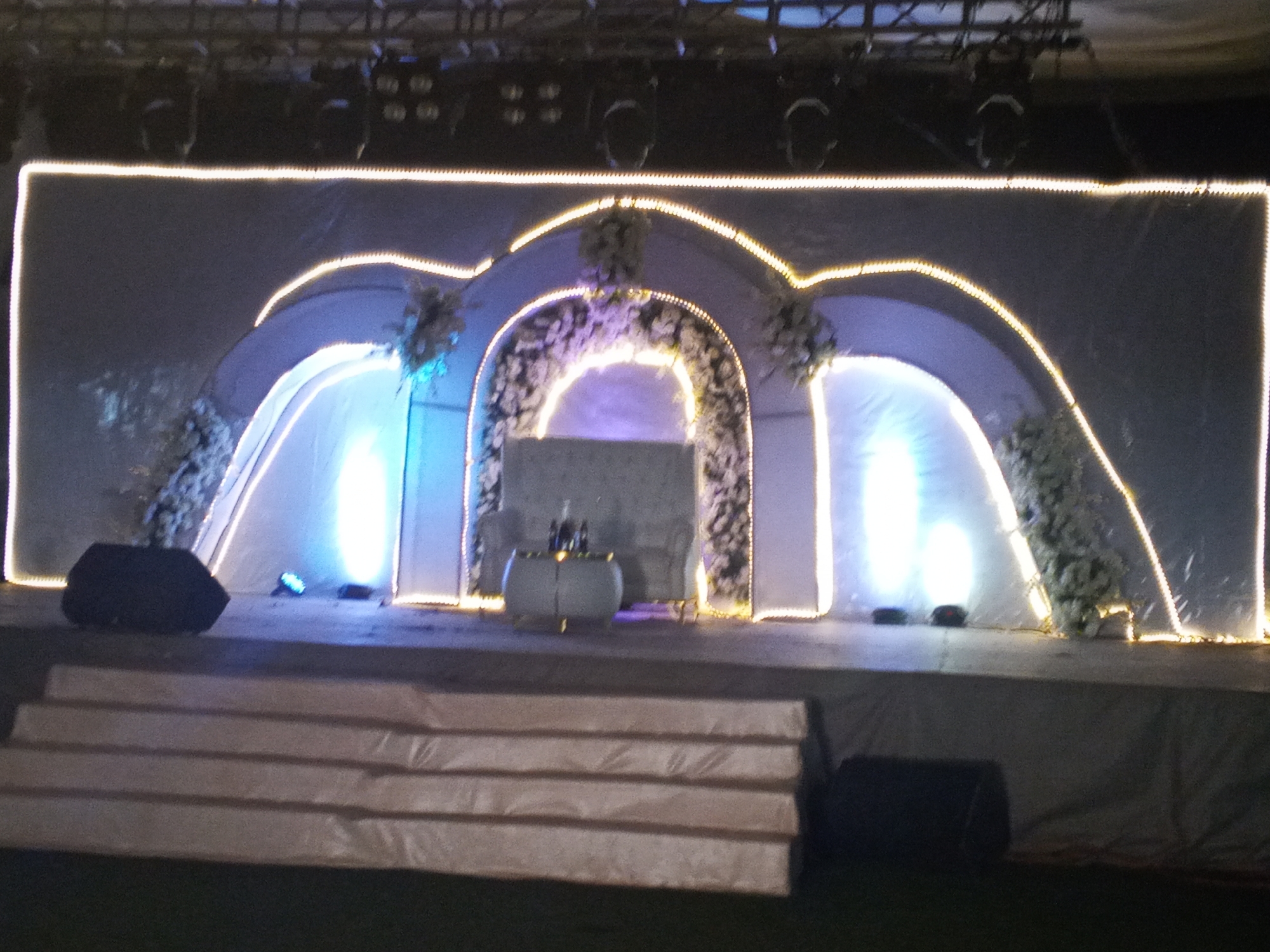 The image shows a stage setup, likely for an event, in what appears to be Jos, Nigeria. The stage is decorated with white fabric backdrops, arches outlined with yellow string lights, and floral arrangements. A round table sits center stage with what appears to be bottles on top. The stage is accessed by a set of covered stairs. Two black speakers are placed on the stage and at the base of the stairs. Above the stage are lights mounted on a truss system.