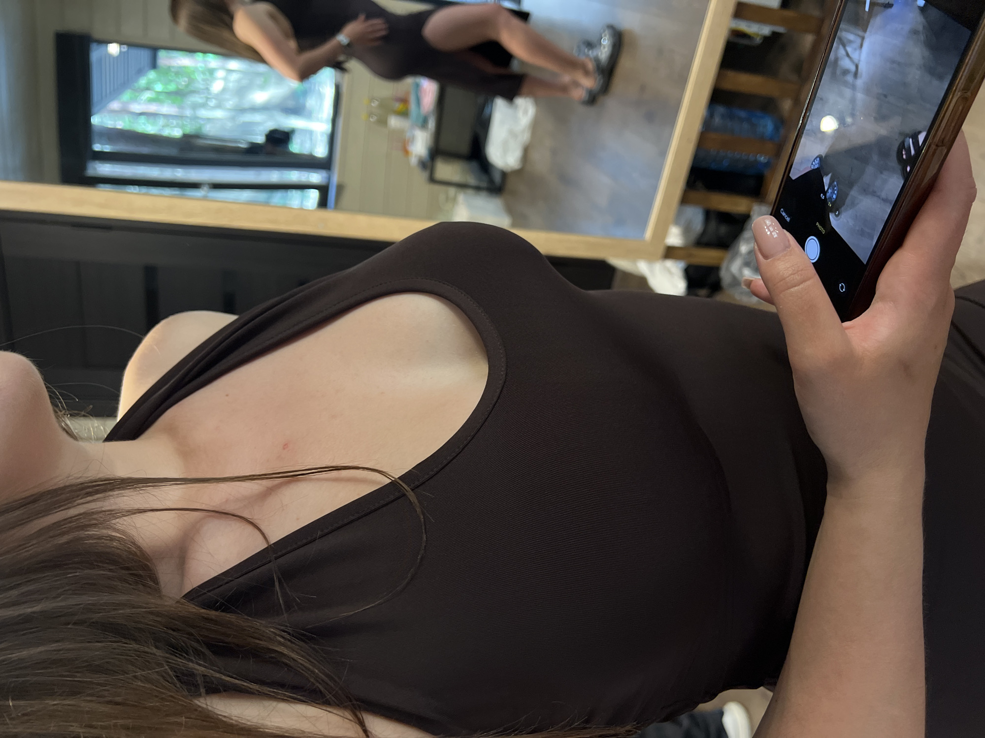 The image shows a close-up of a person's chest and shoulder area, wearing a dark brown sleeveless dress. A hand is visible in the lower right corner, holding a smartphone that is pointed towards a mirror. In the mirror's reflection, a second person is visible, also wearing a dark dress with a high slit on the side. The background in the mirror appears to be an indoor space with wooden walls and a window showing trees outside, possibly indicating a location outside of a major city. The image does not provide any specific details about the city of Ivachiv Horishnii, Ukraine.