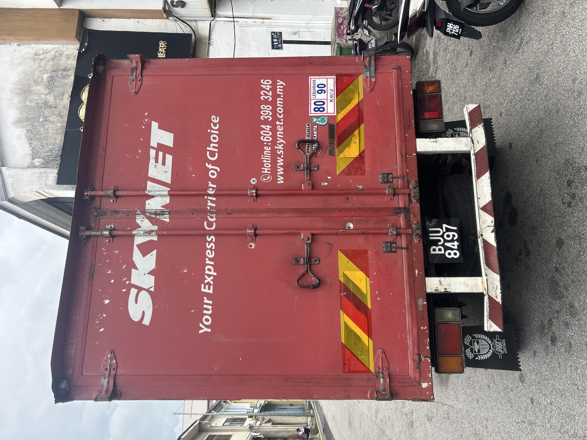 The image shows the rear of a red Skynet delivery truck parked on a street in George Town, Malaysia. The truck has the Skynet logo and the text "Your Express Carrier of Choice" printed on its back doors. Below this text, a phone number "604 398 3246" and a website "www.skynet.com.my" are visible. A speed limit sign indicating 80 and 90 Km/J is also affixed to the truck, along with a Petronas logo. The license plate of the truck reads "BJU 8497".
In the foreground, two motorcycles are parked, one with a license plate "PF1 4518" and another with "PNH 7126". In the background, traditional shophouses line the street, some with faded paint and ornate architectural details. A person can be seen sitting on a step on the left side of the street. The sky is overcast. The scene depicts a typical street view in a historic urban area of George Town, a UNESCO World Heritage Site known for its well-preserved colonial architecture and vibrant street life.