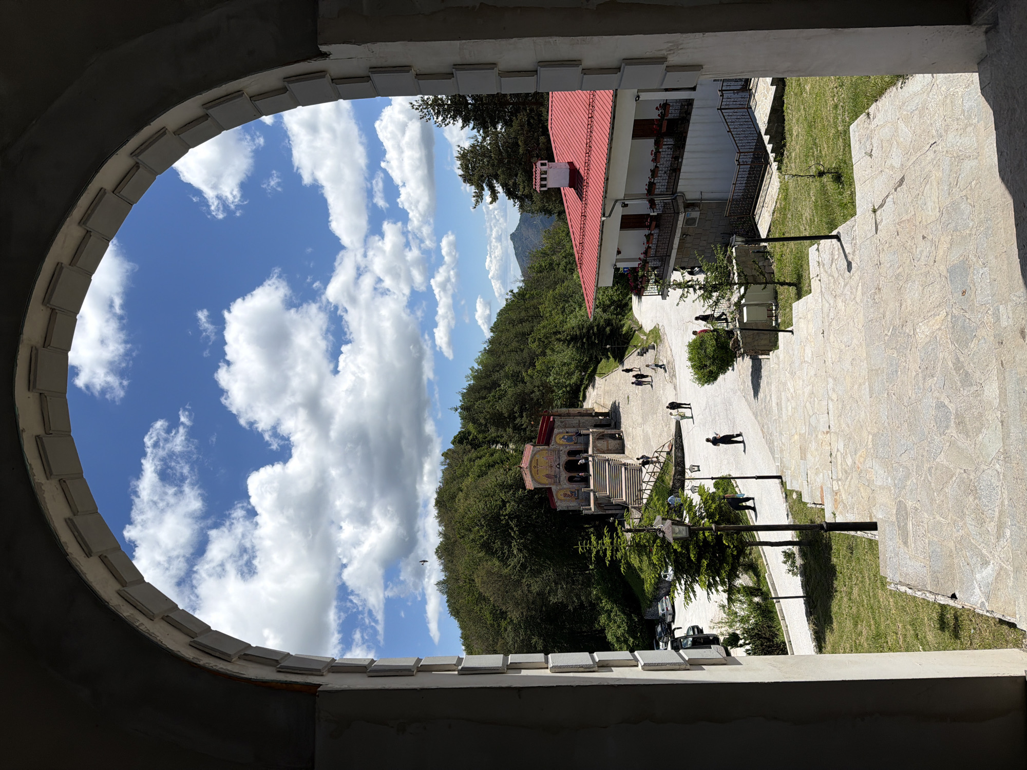 Framed by a large, unfinished concrete archway, the image looks out onto a sunny, mountainous landscape in Mostovo, Bulgaria. The scene depicts a path leading towards a small, stone church with a red-tiled roof and ornate facade, nestled amongst lush green trees. To the right, a white building with a prominent red roof and balconies laden with flowers is situated. Several individuals are visible walking on the path and near the church. The sky is a vibrant blue, dotted with fluffy white clouds. The overall atmosphere is peaceful and serene, with a sense of religious devotion and natural beauty.