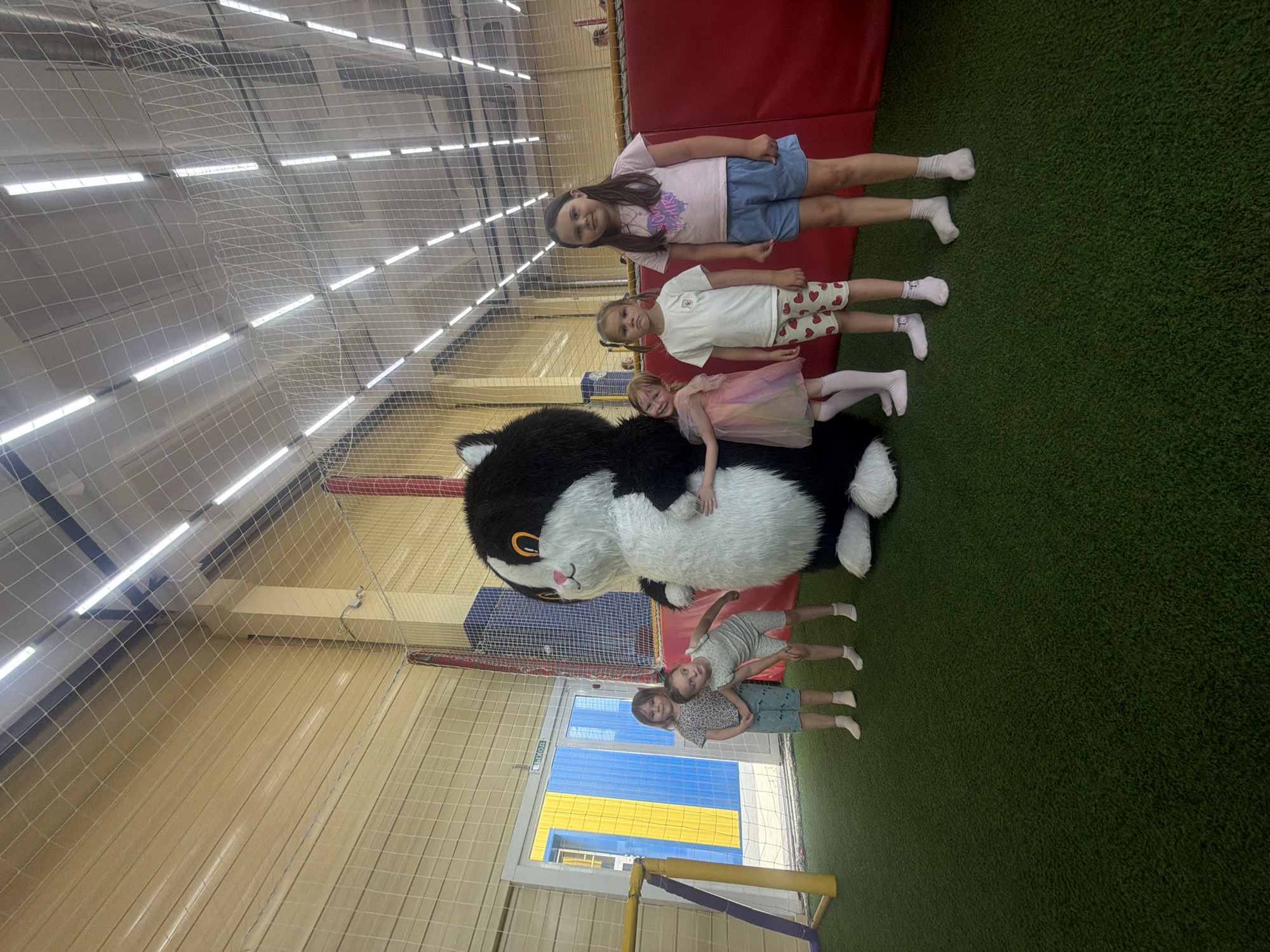 Five children and a mascot costume are posing for a photograph in an indoor playground in Chaikovsky, Russia. The mascot is a large, black and white cat costume with large, round orange eyes and a fluffy white belly and tail. It is standing in the center of the image, with its arms around two young girls. The children are standing in a line to the right of the mascot. The youngest child, wearing a pink and white dress, is leaning into the mascot. The next child, wearing a white t-shirt and heart-patterned shorts, stands with a neutral expression. The third child, wearing a pink t-shirt and blue shorts, stands with a slight smile. The two oldest children are to the far left, closer to a large blue and yellow doorway, with one looking towards the camera and the other glancing away. The playground has a green artificial grass floor and a red padded wall in the background. The ceiling is high and features a netting structure with multiple fluorescent lights. The doorway behind the children appears to be an exit, indicated by the Russian word "ВЫХОД" (VYKHOD) meaning "EXIT". The overall scene suggests a recreational environment for children.