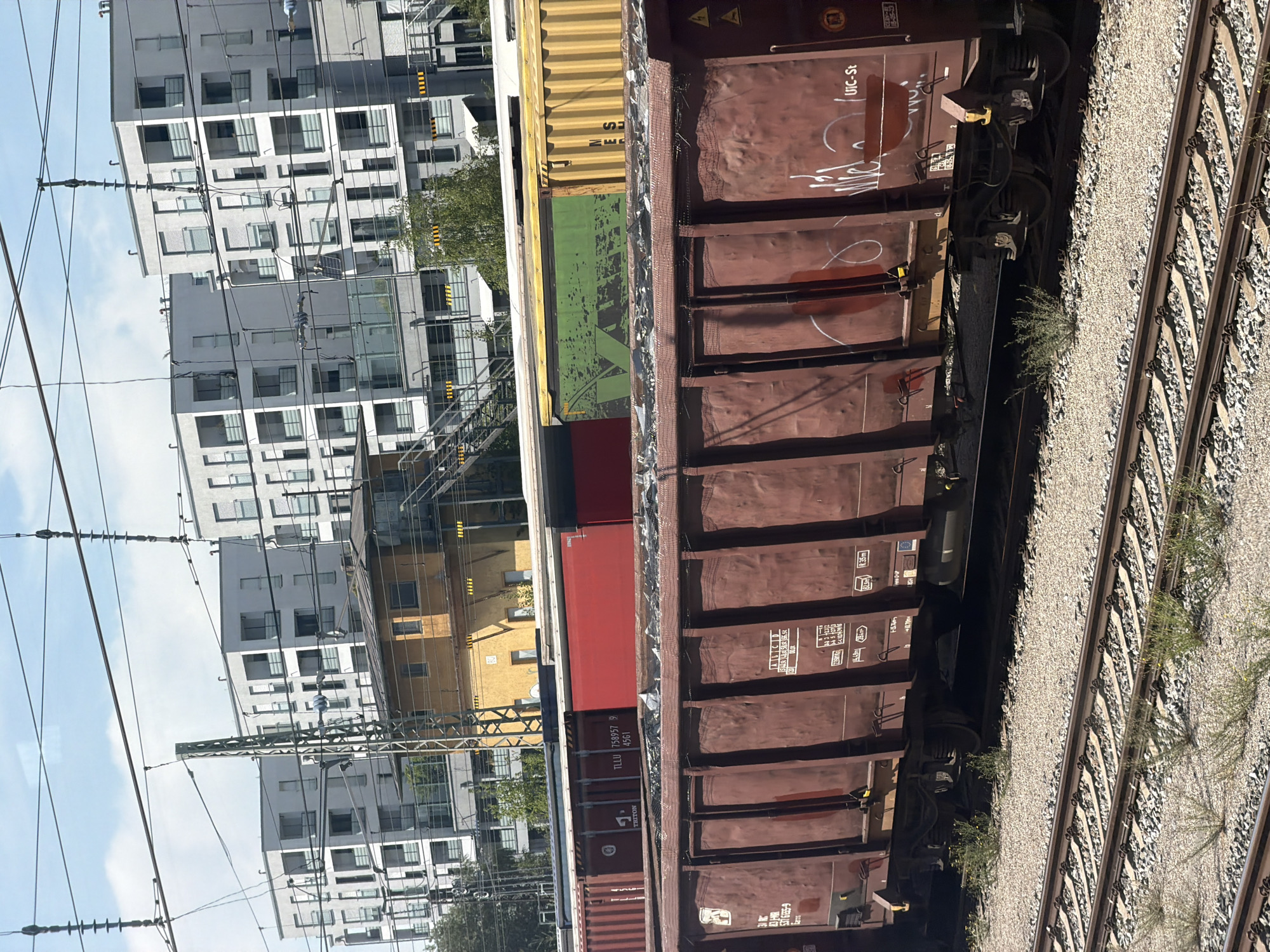 The image depicts a railway yard in Munich, Germany, with a focus on freight trains. In the foreground, a large, dark red freight car is visible, filled with what appears to be debris. The car has markings on its side, including "UIC-St" and some graffiti. Behind this car, a row of other freight cars can be seen, including one with a red container, another with a green container, and a yellow container. In the background, a complex of modern, white apartment buildings with balconies dominates the skyline. Between the apartment buildings and the railway yard, a smaller, yellowish building with a distinctive architectural style stands out. Overhead, electrical wires for the trains are strung across the frame, supported by metal poles. The scene is bathed in daylight under a partly cloudy sky.