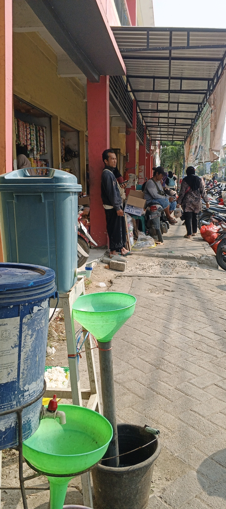 In Sumberwudi, Indonesia, a street-level view captures a scene along a building's facade, likely a storefront or market. A weathered blue cylindrical container, possibly a water tank or large barrel, stands prominently on the left, displaying some label text. Adjacent to it, a green funnel is suspended over a pipe, with a red tap connected to the blue container dripping water into the funnel, suggesting a makeshift water dispensing station. Below the funnel, a dark-colored bucket sits on the paved ground.
A man in dark trousers and a grey long-sleeved shirt stands near a red pillar, looking to his right, holding a cigarette and a phone. Further down the covered walkway, another individual sits on a motorcycle, also holding a cigarette and looking down. Behind them, a woman in a patterned headscarf and dark clothing walks away from the camera. The walkway is lined with parked motorcycles. The building has yellow and red painted sections, with a metal awning overhead supported by black metal beams. The ground is paved with interlocking bricks. The overall impression is of a functional, everyday scene in a semi-urban Indonesian setting.