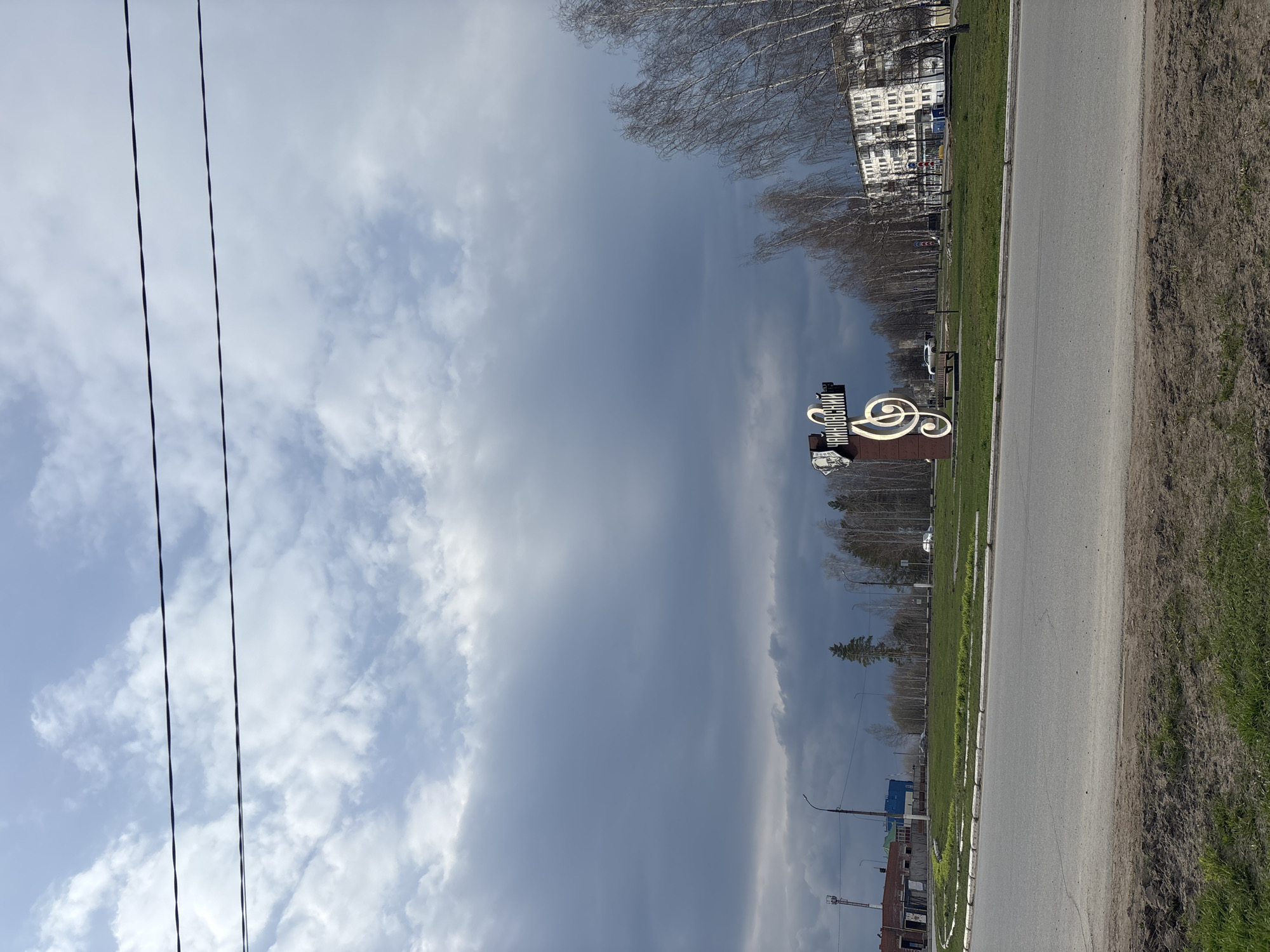 This daytime, landscape-oriented photograph captures a street scene in Chaikovsky, Russia. The upper portion of the image is dominated by a partially overcast sky, featuring a mix of bright, scattered clouds and a larger, darker gray cloud formation. Two dark power lines run vertically across the left side of the sky.
In the middle ground, a paved road with a white edge stripe extends into the distance. To the immediate right of the road, there is a strip of green grass, which transitions into an area of bare, brown earth. To the left of the road lies a broader expanse of green grass.
A notable decorative sign is positioned on the left side of the road in the foreground. This sign features a prominent, stylized musical treble clef in light tones, alongside another curved musical motif. A rectangular element attached to the sign displays the Cyrillic text "ЧАЙКОВСКИЙ" (Chaikovsky), referencing the city's name and its connection to the composer Pyotr Ilyich Tchaikovsky.
In the background, a line of deciduous trees with bare branches suggests either early spring or late autumn. Behind these trees, several multi-story buildings, including a distinct white structure, are visible, hinting at an urban or suburban setting. No people or specific activities are present in the static scene. The weather appears temperate with significant cloud cover.