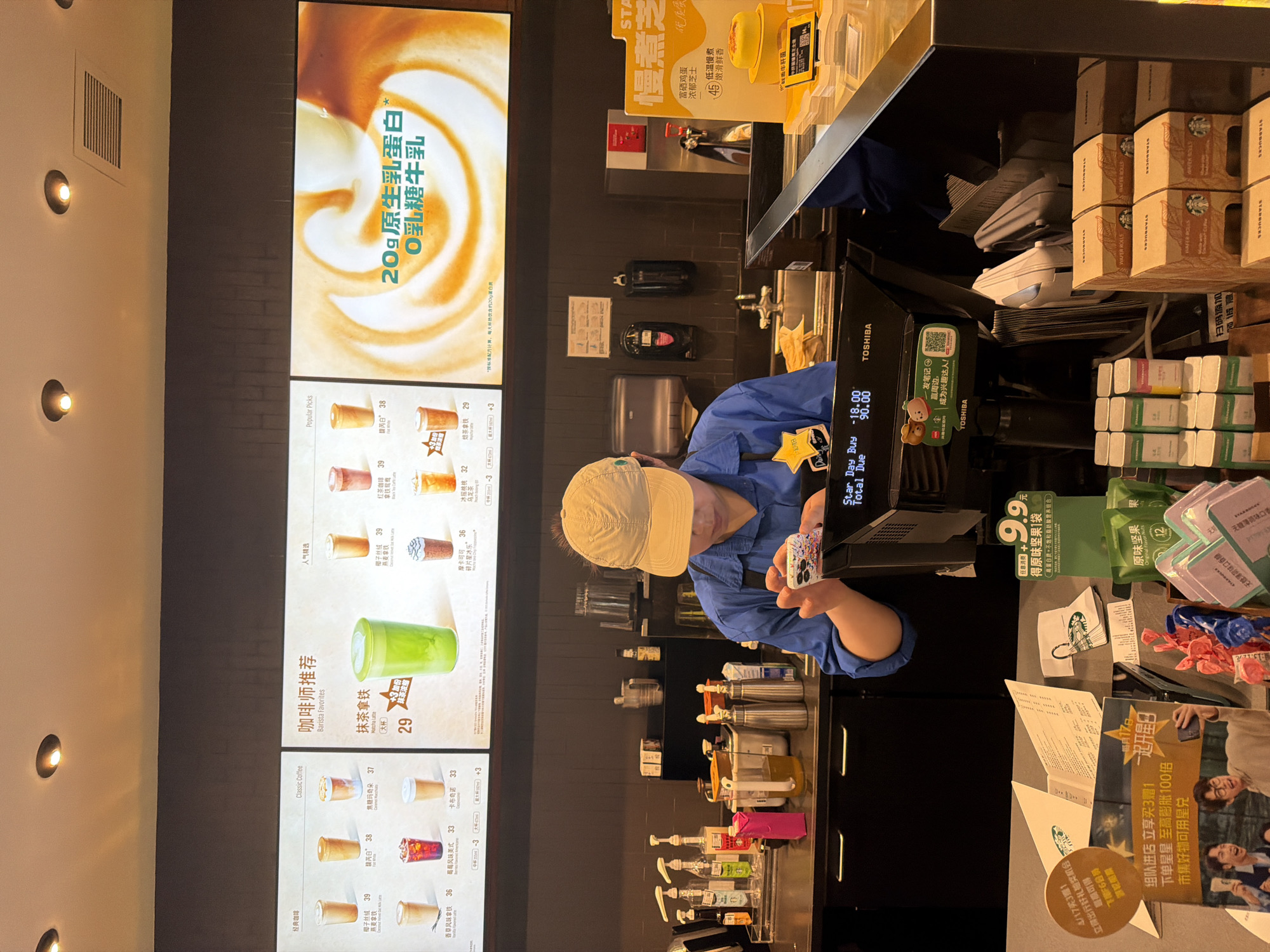 The photo shows the interior of a Starbucks store, likely located in Yuhuatai District, China, based on the presence of Chinese characters and references to promotions specific to that region. A Starbucks employee wearing a blue uniform and a khaki cap is holding a smartphone with a brightly colored case near the point-of-sale terminal.
To the left of the employee is a digital menu board displaying a variety of drinks with prices listed in Chinese currency. The menu includes classic coffees, barista favorites like matcha lattes, and popular picks such as coconut velvet and oat lattes. Advertisements for promotional offers are also present on the board, one of which highlights a product with 20 grams of native milk protein.
In front of the employee are the payment terminal, and multiple stacks of Starbucks-branded wafer roll boxes. Various packaged food items, including packets of "original flavor nuts" and "sugar-free mint flavor gum" are visible to the right of the terminal. Promotional material advertising a "Buy 3 get 1 free" offer is also displayed.