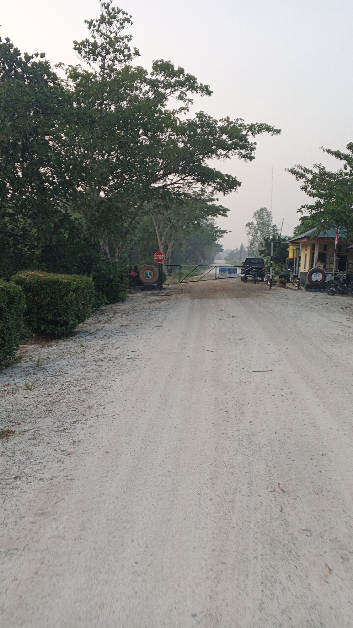 The image shows a dirt road leading to a closed gate. On the left side of the road, there are neatly trimmed bushes and trees. A red stop sign is visible near the gate, along with a circular emblem. On the right side of the road, there is a building with a yellow facade and a red roof. A black vehicle is parked near the gate, and several motorcycles are also present. The sky is overcast, and the overall impression is one of a rural or semi-rural setting. The location is identified as Merempan Hulu, Indonesia.