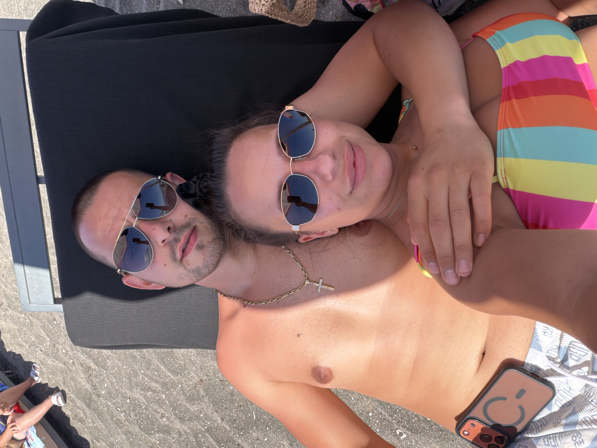 A man and woman are lying on lounge chairs on a beach. The man is shirtless, wearing sunglasses, a gold chain with a cross pendant, and patterned swim trunks. The woman is wearing sunglasses and a colorful, striped bikini. They are both looking at the camera and embracing. A phone with a brown case is on the sand next to the man. In the background, another person is visible sitting on a lounge chair. The beach is covered in dark sand and small pebbles. The overall scene suggests a sunny day at the beach. The location is identified as Burgas, Bulgaria.