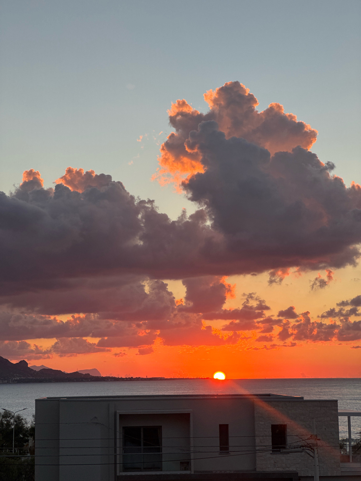 The image captures a vibrant sunset over the sea, with dramatic clouds tinged with orange and pink. In the foreground, the top of a modern building with a white facade and dark-framed windows is visible. Beyond the building, the calm sea stretches out to the horizon, where a sliver of the sun is just dipping below. Distant, dark mountains form a silhouette against the fiery sky. The location is identified as Isola delle Femmine, Italy, a coastal town known for its natural beauty. The scene is peaceful and serene, with no people or specific actions depicted.
