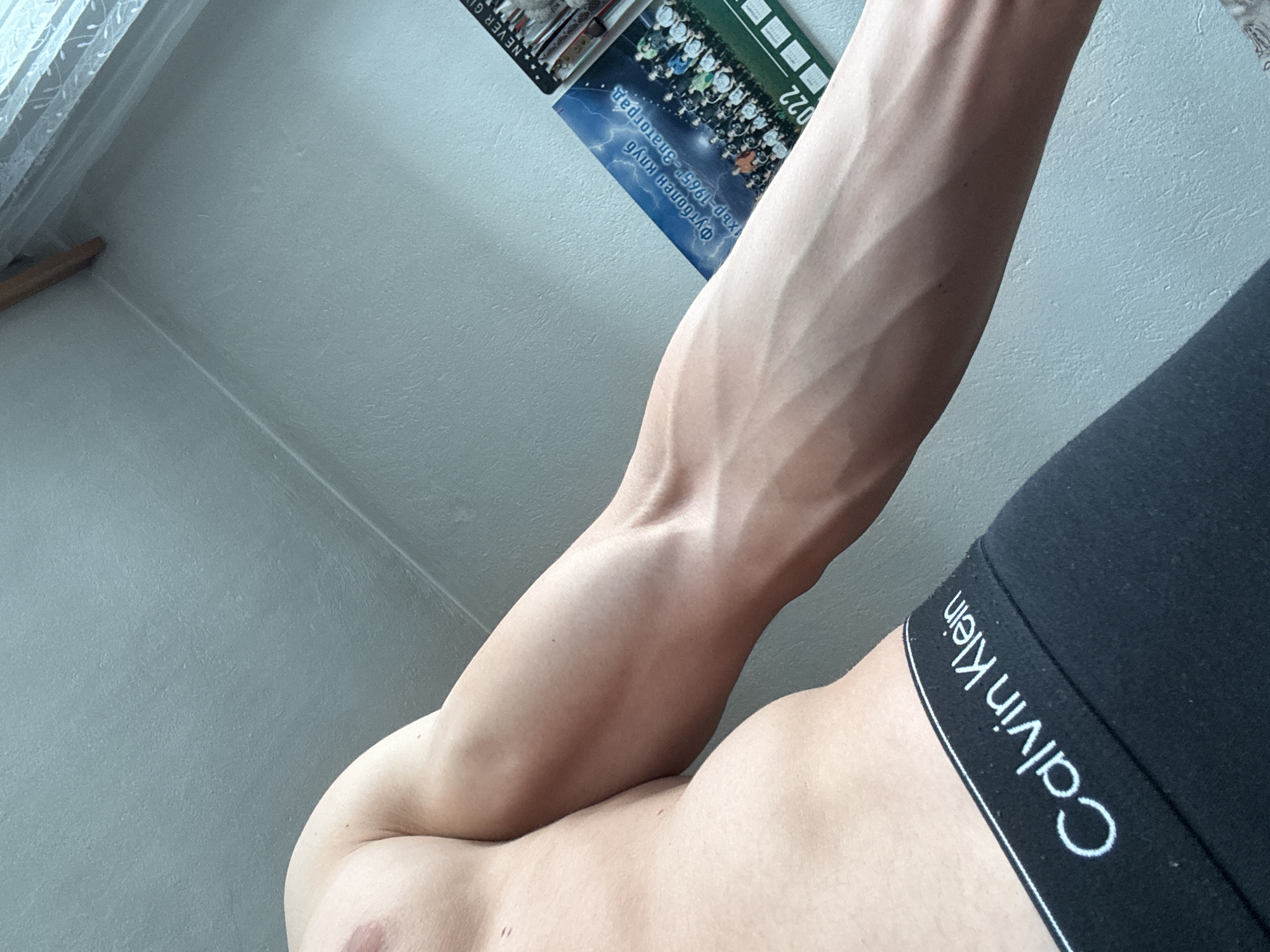 The image shows a person's arm and part of their torso, turned at an angle. The arm is flexed, revealing prominent veins. The person is wearing black Calvin Klein underwear. A calendar hangs on the wall behind the arm. The calendar features the words "Футболен Клуб ХБР-1965г. Златоград" ("Football Club HBR-1965 Zlatograd"), indicating the location is Zlatograd, Bulgaria. A portion of the calendar shows an image of what appears to be a football team. A window and curtain are visible in the top left corner.