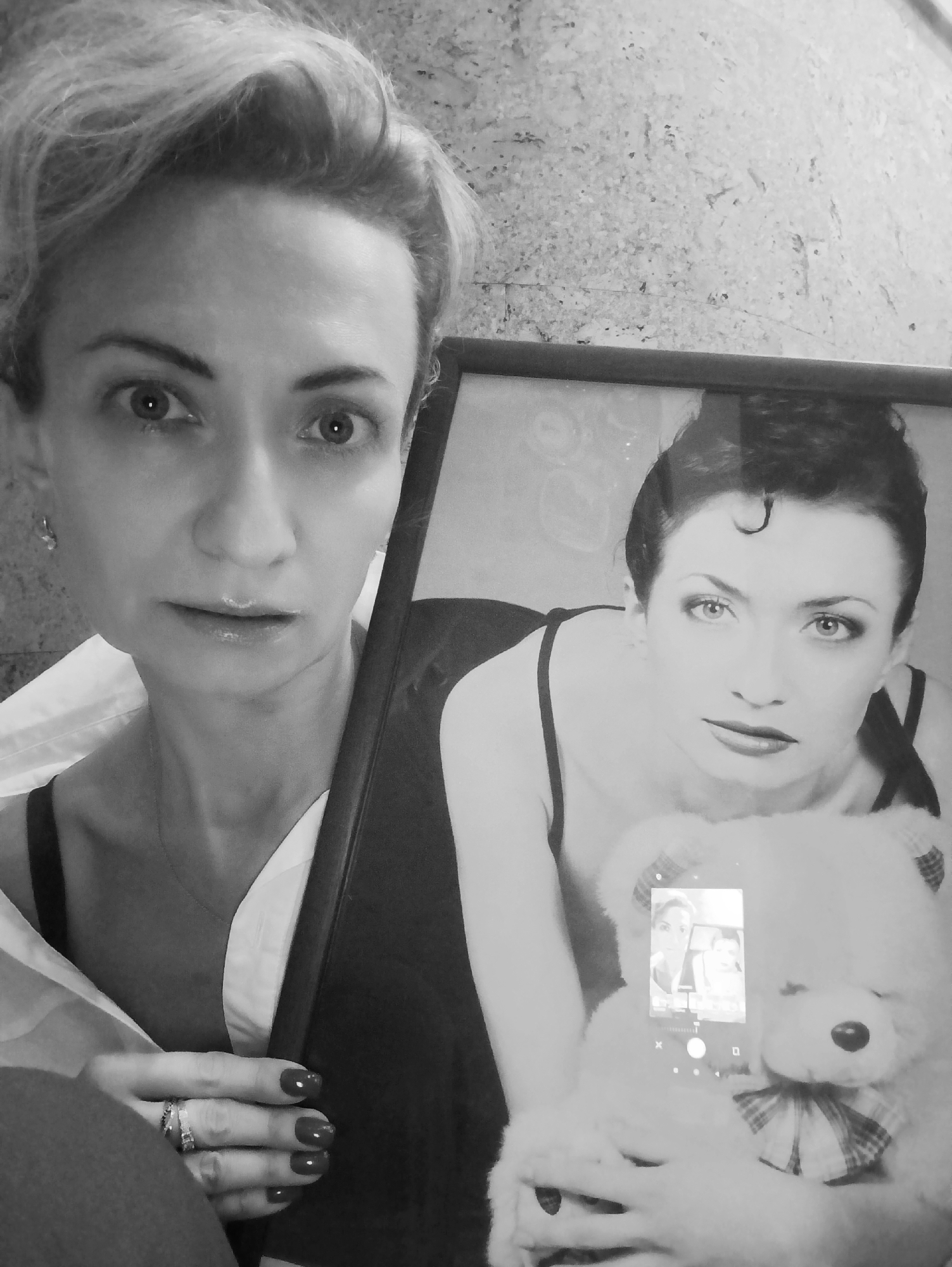 A black and white selfie of a woman holding a framed photograph and a large teddy bear. The woman, who appears to be in her late 30s or early 40s, has short, light-colored hair, styled upwards. She is wearing a white button-down shirt with the collar open, revealing a dark bra underneath. Her left hand, adorned with rings and dark nail polish, is visible in the foreground, holding the framed photo. The framed photograph shows a younger woman with dark hair, styled up with a curl on her forehead, wearing a dark camisole. This woman in the photo is also holding a large white teddy bear with a plaid bow. The background of the selfie appears to be an indoor setting with a textured wall. There is no discernible information in the image that would indicate a specific location within Moscow, Russia.