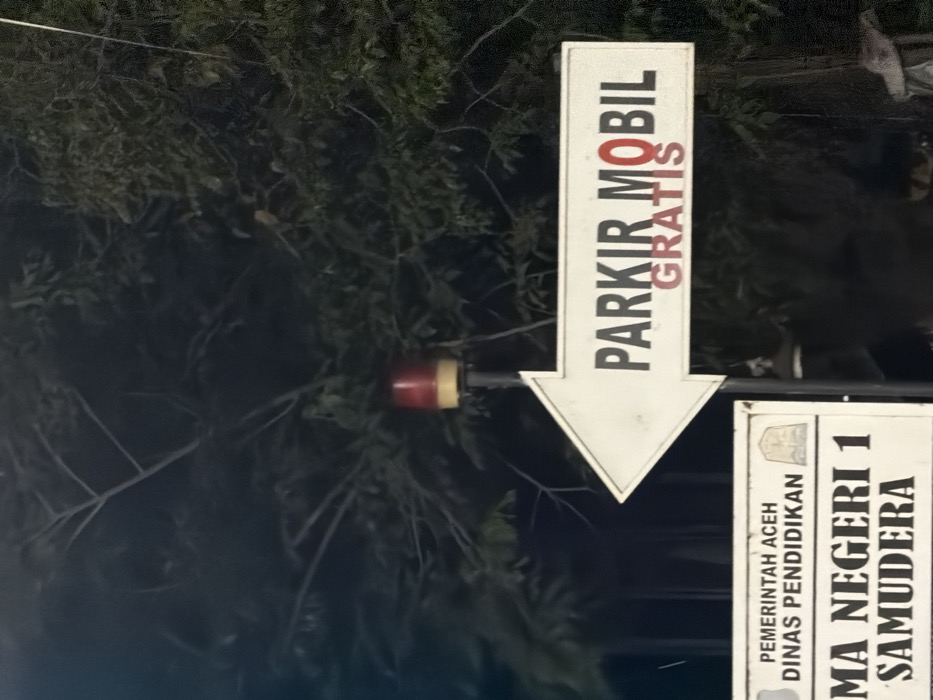 The image displays a nighttime or dusk scene featuring two informational signs and a warning light against a dense, dark green foliage background. A prominent white arrow-shaped sign points left, bearing the Indonesian text "PARKIR MOBIL" (Car Parking), with the word "GRATIS" (Free) overlaid in red, partially obscuring the letter 'O' in "MOBIL." Directly above this sign, a red and amber/yellow rotating beacon or warning light is mounted on a pole. Below the arrow sign, a rectangular white sign is visible, displaying "PEMERINTAH ACEH DINAS PENDIDIKAN" (Government of Aceh, Education Department) and "MA NEGERI 1 SAMUDERA" (Public Senior High School 1 Samudera), accompanied by an institutional emblem. The vegetation in the background consists of trees or bushes, indicating an outdoor, possibly semi-urban or rural setting. No individuals or active interactions are present. The signs confirm the location is at or near Public Senior High School 1 Samudera, situated in the Samudera district of North Aceh Regency, Aceh Province, Indonesia, which includes the specified area of Beurandang.