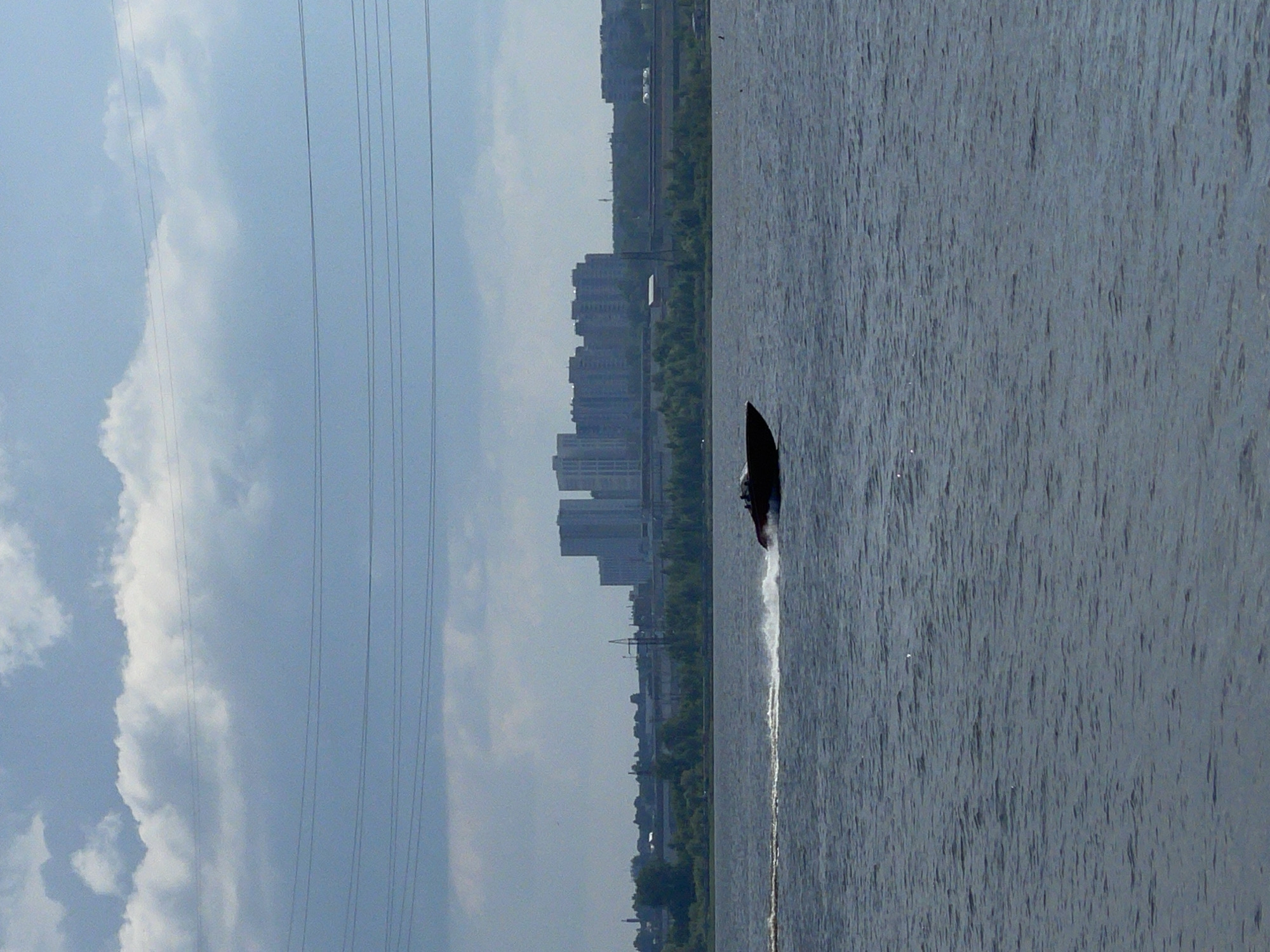 The image depicts a wide, rippled body of water, likely a river or lake, with a dark-colored speedboat cutting through the water from left to right, leaving a white spray wake behind it. In the background, a distant cityscape is visible, characterized by numerous multi-story buildings, suggesting an urban environment. A line of trees separates the water from the buildings. Above the cityscape, a network of power lines stretches across the frame, silhouetted against a cloudy sky. The overall scene suggests a view of a city on a somewhat overcast day, with a boat actively navigating the waterway. The location is identified as Izhevsk, Russia, though no specific landmarks within the city are clearly identifiable from this image.
