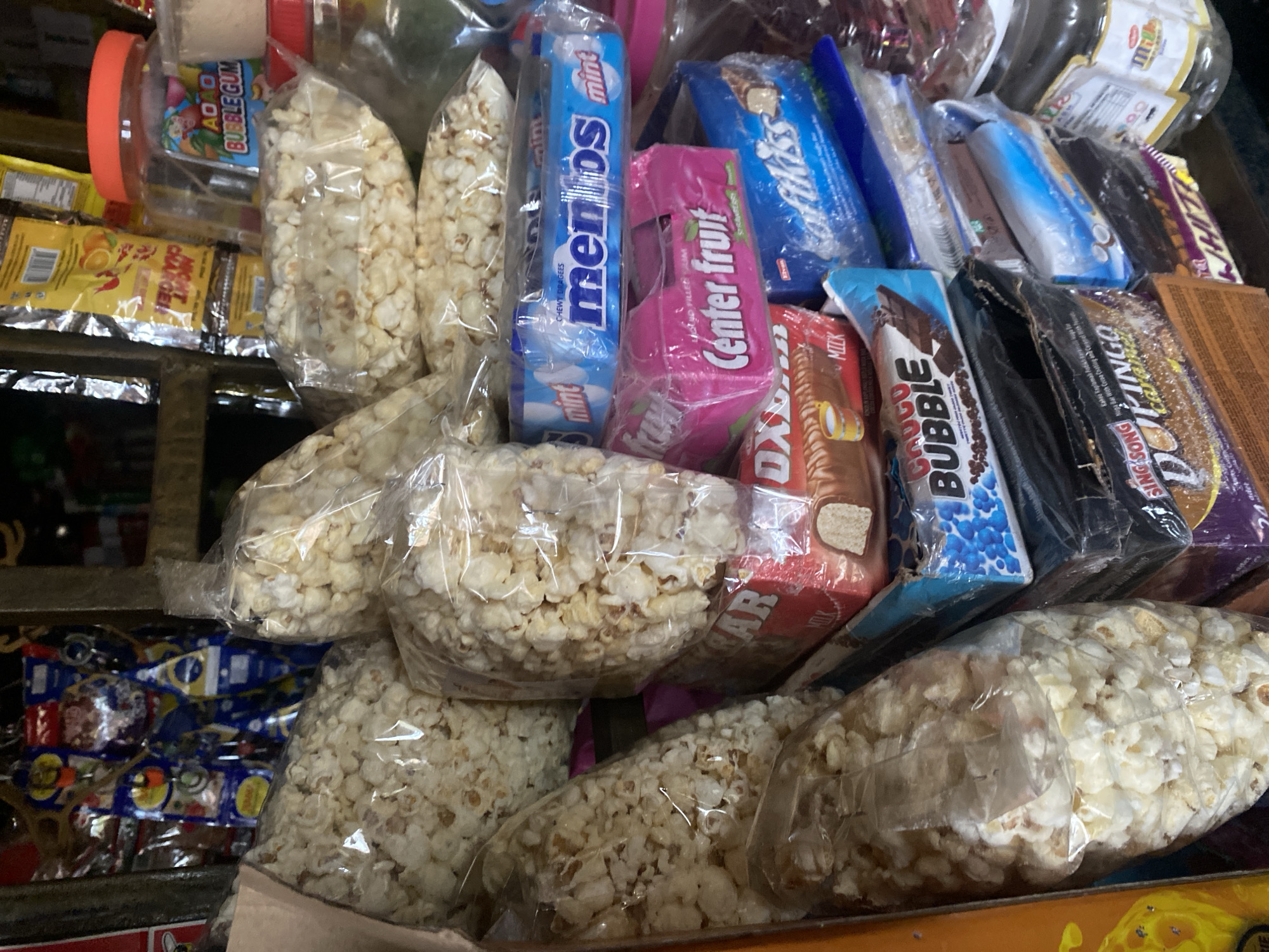 The image depicts a close-up view of a stall in a Nigerian market, specifically in Lafia, Nigeria. The stall is filled with a variety of snacks and candies, primarily packaged items. Prominently displayed are several large, clear plastic bags filled with popcorn. Other items visible include packs of Mentos, Center Fruit, a chocolate bar labeled "CHOCO BUBBLE," and a candy bar with "SING SONG" and "DOKINGO" visible on its packaging. A jar with an orange lid, containing what appears to be candy, is also present. The background shows more packaged goods and the general clutter of a market stall. The lighting is somewhat dim, suggesting an indoor or shaded market setting. There are no people visible in the frame.