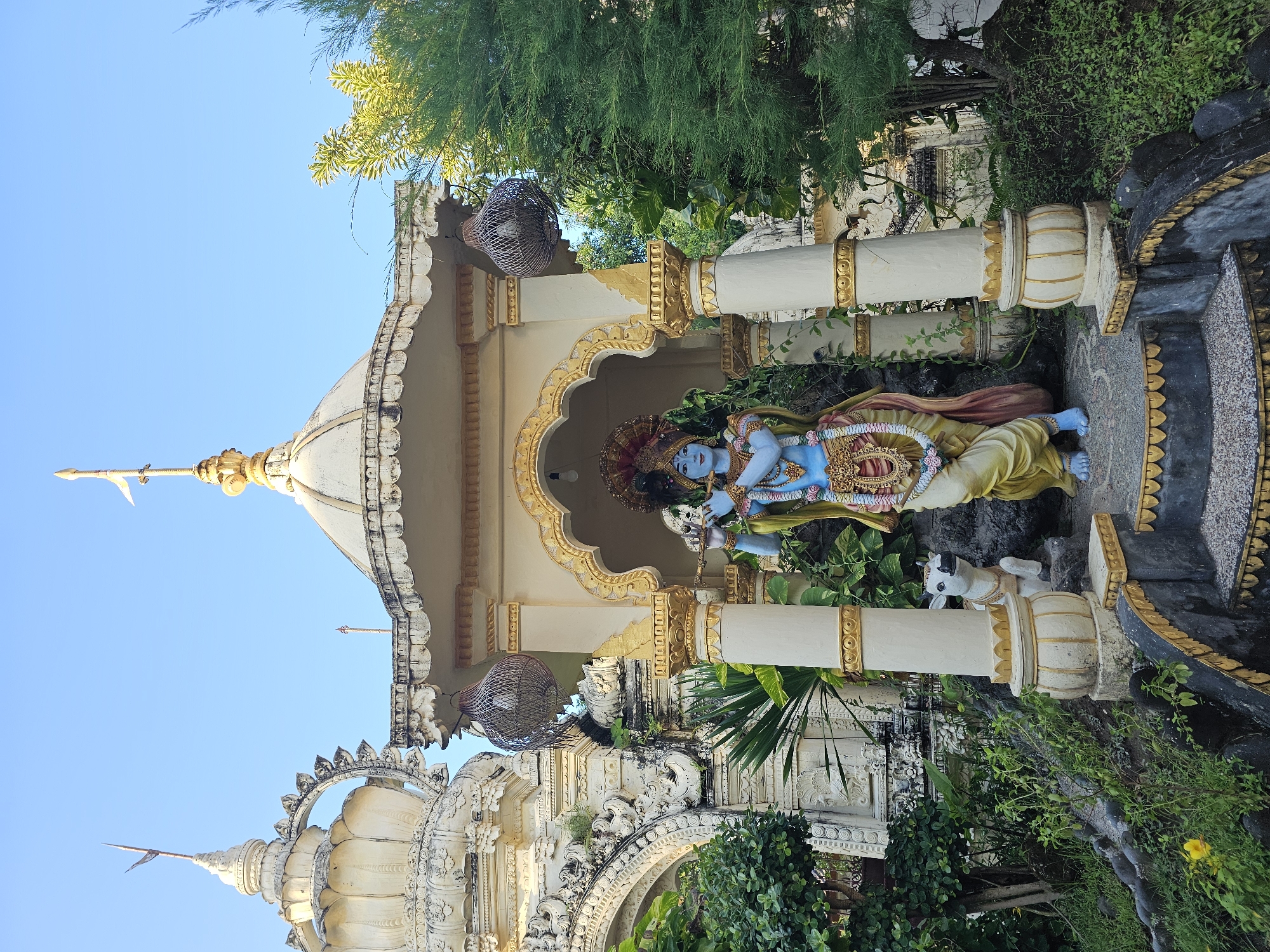 The image depicts a statue of Krishna playing the flute within a temple structure. The statue is positioned between two white and gold columns. A small white statue of a cow sits beside Krishna. The temple building features intricate carvings and is adorned with plant life. The sky is clear and blue. Based on visual cues and cultural indicators, the location is likely a Hindu temple, possibly in Denpasar, Indonesia.