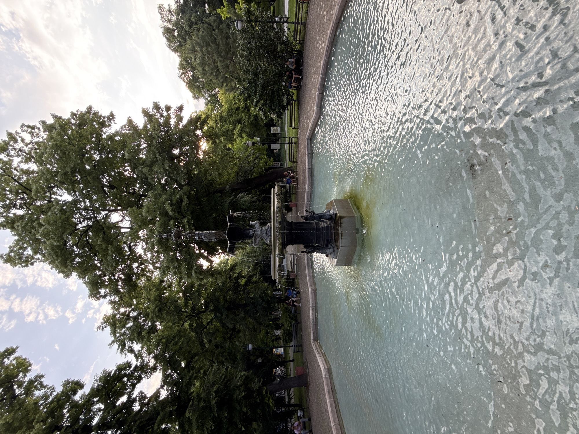 This image captures the "Perla" (Pearl) fountain situated within the Tsar Simeon Garden in Plovdiv, Bulgaria. The central focus is a large, multi-tiered classical fountain constructed from dark metal or bronze. The fountain's uppermost tier features a statue of a standing female figure, while water cascades from various levels into a broad, octagonal basin at ground level. The water in the basin is a clear, light blue-green, displaying ripples and reflections.
The fountain is encircled by a paved pathway of light-colored, possibly brick or cobblestone, material. Beyond the pathway, the park environment is characterized by dense green foliage from numerous mature trees of varying heights, creating a shaded canopy. Several dark-colored park benches are positioned along these pathways beneath the trees.
Multiple individuals are observed in the scene. On the left side of the fountain, two people are seated on a bench, one wearing a pink shirt. On the right side, a group of at least three individuals is seated on another bench further back. Dark, ornate street lamps are visible at various points within the tree line.
The sky above is a pale blue adorned with scattered, wispy white clouds, indicative of late afternoon or early evening. Soft, warm light filters through the trees from the left, illuminating parts of the scene and reflecting off the water's surface.