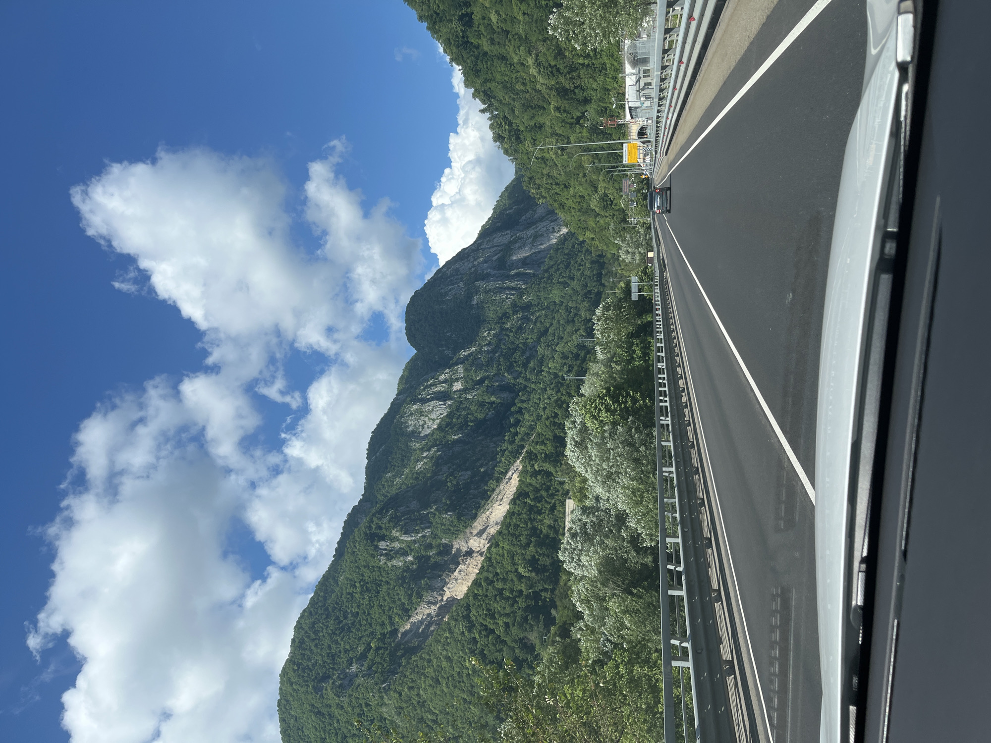 The image depicts a road trip through a mountainous region with a clear blue sky and scattered white clouds. The road is paved with white lane markings and flanked by metal guardrails. On either side of the road, lush green trees and steep, rocky mountains rise dramatically. One mountain face shows a visible landslide scar. In the distance, a tunnel entrance is visible on the right side of the road, and a few cars are present on the highway. The viewpoint is from inside a vehicle, looking forward. The location is not Monastyr, Russia, as this image depicts a mountainous landscape inconsistent with the known geography of Monastyr.