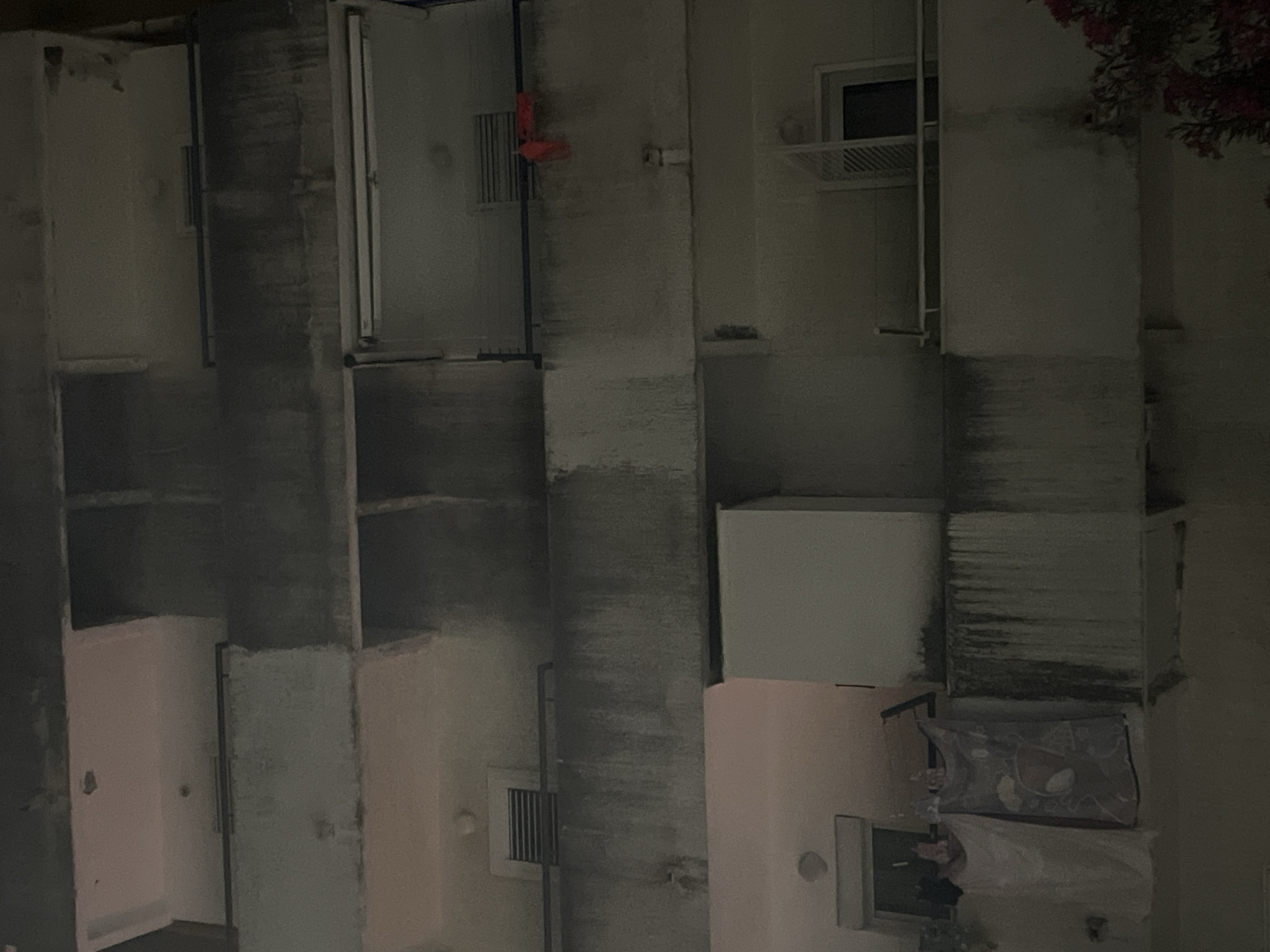 The image depicts a nighttime view of the exterior of a multi-story building, likely an apartment complex, in Budva, Montenegro. The architecture features multiple balconies, some with railings, and visible windows and vents. The building's facade appears to be made of concrete and shows signs of wear, with staining and discoloration on some of the balconies.
On one of the lower balconies, laundry is hung out to dry. A white sheet or towel is draped over a railing, and a patterned blanket or towel hangs next to it. A person, partially obscured, is visible near the window of this balcony.
On a balcony on a higher floor, a piece of bright orange fabric is draped over the railing. Another balcony on a higher level has its shutters partially open, revealing a dark interior.
The overall lighting suggests it is nighttime, with some interior lights or ambient light illuminating parts of the building. In the lower right corner of the image, a cluster of pink and red flowers is visible, suggesting the presence of vegetation near the building. The scene is presented in a somewhat desaturated and grainy manner, consistent with a low-light photograph.