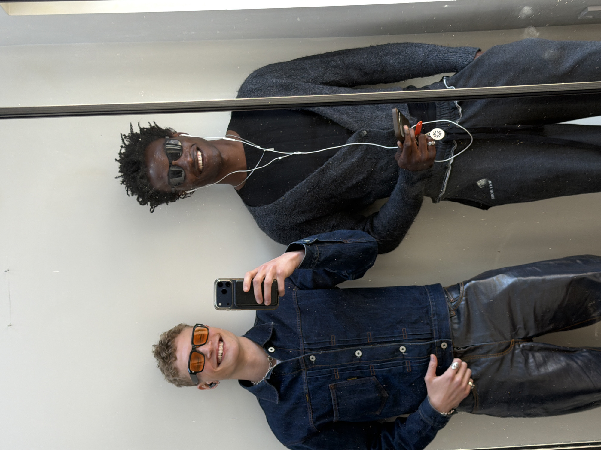 Two men are captured in what appears to be a mirror selfie or reflection, seen within an indoor setting in London, United Kingdom. Both subjects are smiling broadly, conveying a happy and relaxed mood. The image is brightly lit, suggesting daytime.
The man on the left has dark skin and black, textured hair, and is wearing dark rectangular sunglasses. He sports a black t-shirt under a dark grey, textured cardigan and dark grey sweatpants. White wired earphones are visible, connecting to a dark-colored smartphone he holds in his right hand. He also holds a small white circular object with a red clip attached. Several silver rings adorn his fingers. On his sweatpants, partially visible due to reflection, is text that reads "SHOM YAW HOME."
The man on the right has lighter skin and light brown curly hair. He wears orange-tinted sunglasses, a dark blue denim jacket with contrast stitching and light buttons, and dark, glossy leather-like pants. He holds a dark-colored smartphone in his left hand, and silver rings are visible on his right hand.
The background is a plain light grey or off-white, with a thin, dark horizontal bar traversing the scene, possibly a structural element or part of the mirror frame. The overall scene suggests a casual, stylish, and contemporary aesthetic.