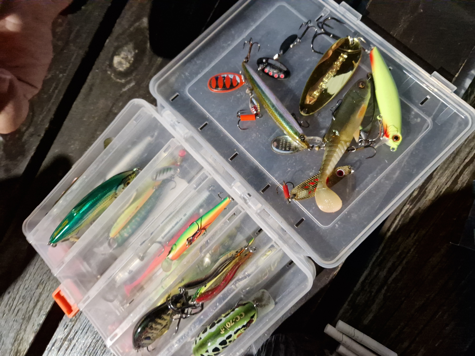 A high-angle medium shot captures a clear plastic tackle box filled with fishing lures. The box is open, with its two sections visible. The left section contains several lures, including a green and black patterned frog lure with "CC-70" printed on it, and a smaller lure with orange and green accents. The right section holds a collection of lures, one with a metallic green body and orange tail, another that is a gold spoon lure, and a pale yellow lure with red markings. Several hooks and a red spinner are also present. The tackle box rests on a dark wooden surface, likely a table, with a few white cylindrical objects, possibly rolled cigarettes, lying to the left of the box. A blurred human hand is visible in the upper right corner of the frame. The scene is set outdoors on a wooden table.