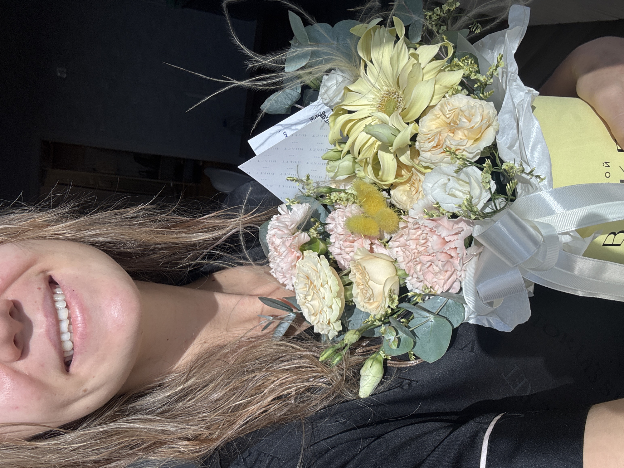 A close-up, slightly rotated image shows a woman's face partially visible on the left side, smiling and exposing her teeth. Her hair, with highlights, flows across the lower part of the image. A bouquet of flowers is positioned above her head. The bouquet contains an arrangement of light yellow and cream-colored flowers, including what appear to be carnations and roses, along with sprigs of greenery and wispy, decorative elements. A white ribbon is tied around the bouquet, which is wrapped in white tissue paper and sits atop a yellow package. The background is predominantly black, providing a stark contrast to the lighter elements of the composition. The location and city are unknown.