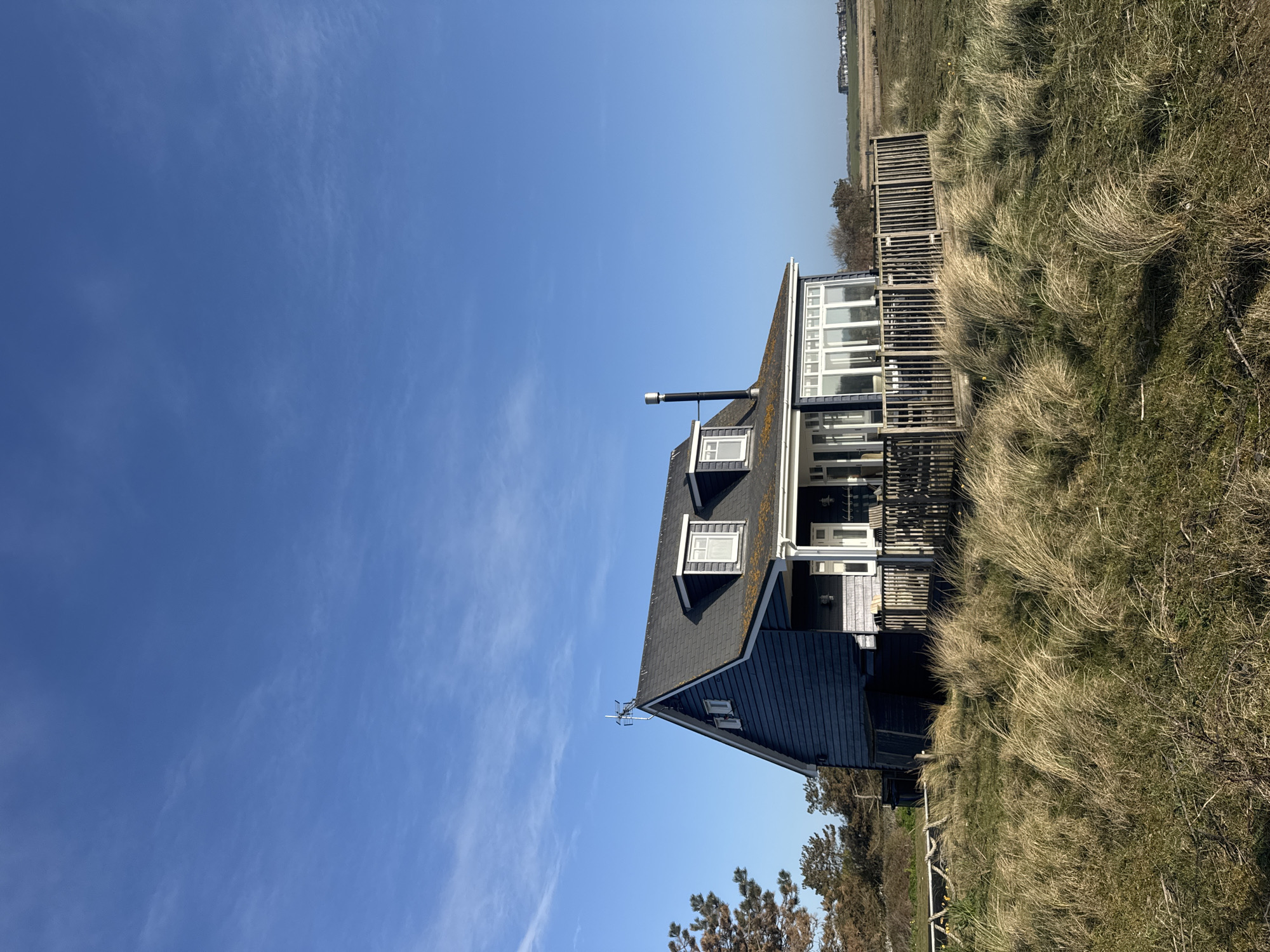 The image depicts a house located in a grassy area with a clear blue sky. The house is dark-colored with a grey roof and white-framed windows. There is a chimney extending from the roof. The house appears to be located in a natural environment, possibly near a beach, based on the surrounding vegetation and open space. There is a wooden boardwalk or pathway extending from the house into the grassy dunes. In the background, some buildings are visible, indicating a nearby settlement. The location is East Suffolk, United Kingdom.