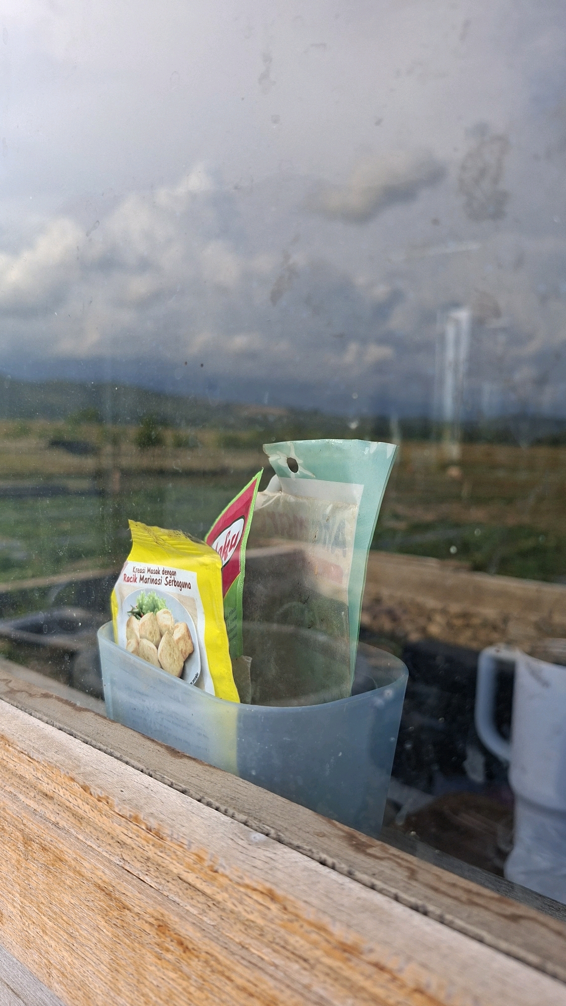 Two small packets of food, one yellow and one green, are placed inside a light blue plastic container. The yellow packet features an image of fried tofu or similar food items and text in Indonesian. Behind these packets, a white mug is visible, reflecting the cloudy sky and a green, grassy landscape. The container and its contents are positioned on a wooden surface, likely a windowsill, with the natural scenery visible through a dirty glass pane. The overall scene suggests an outdoor or semi-outdoor setting, possibly in Gronggrong, Indonesia, given the context.