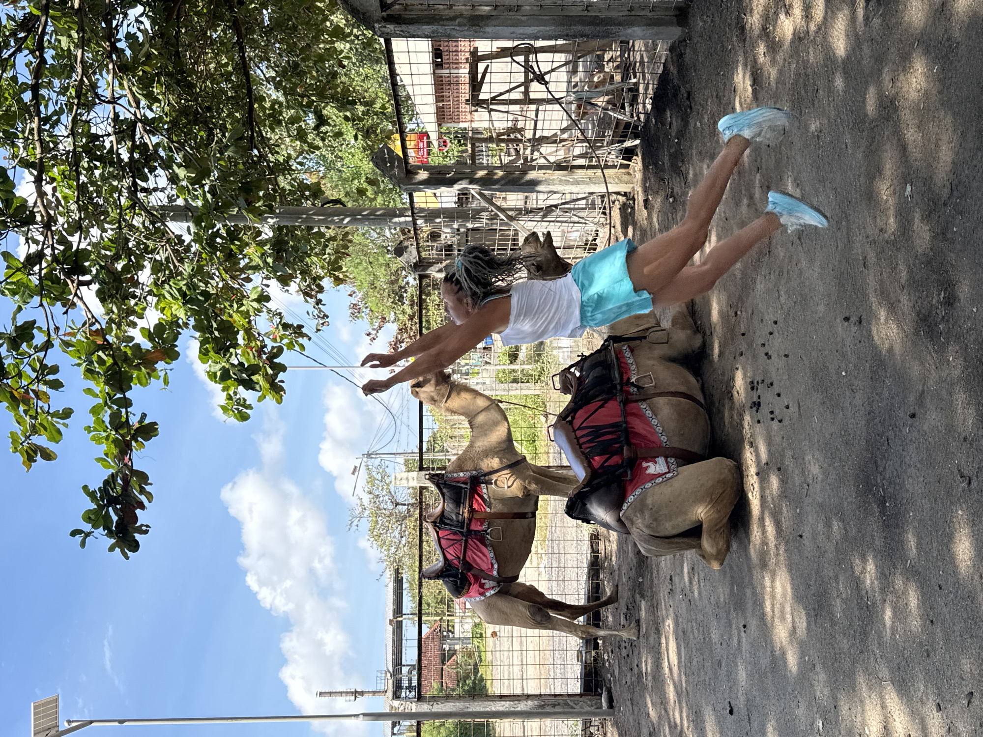 In Kuta, Indonesia, a young woman with braided hair is captured mid-air as she jumps. She is wearing a white tank top and bright turquoise shorts, with light blue athletic shoes. Her arms are raised, and her body is extended upwards. In the background, two camels are present; one is standing with a red and black saddle, while the other is lying down, also equipped with a similar saddle. A wire fence encloses the area, and beyond it, some structures and trees are visible under a bright blue sky with scattered clouds. The ground is dusty and appears to be a dirt or gravel surface.