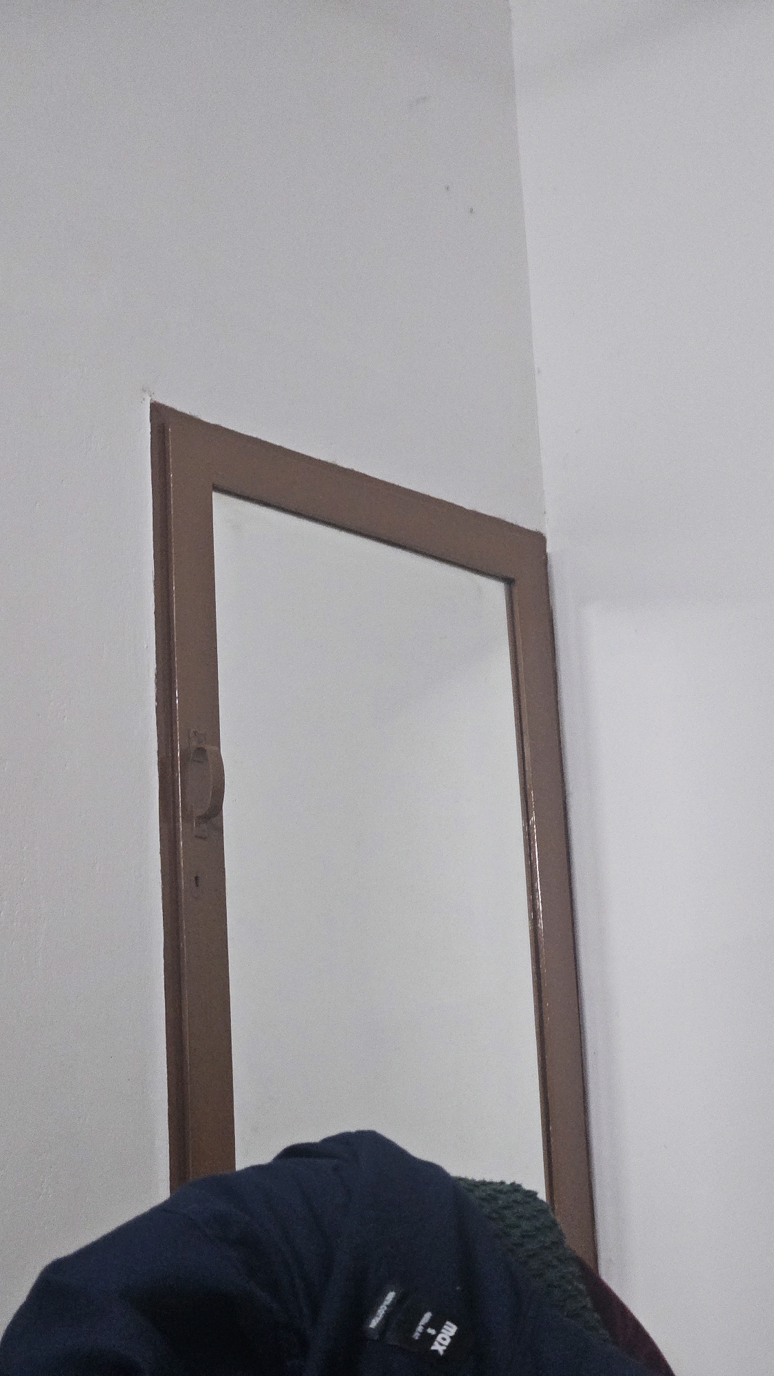 The image shows a close-up of a brown-framed mirror leaning against a white wall, possibly in a room in Choozhattukotta, India. The mirror's surface is blank and reflects the white wall. A dark blue garment with white text is visible in the foreground, partially obscuring the bottom of the mirror. The garment appears to be a piece of clothing, possibly a jacket or shirt, with the text "max" and a logo clearly visible. The environment appears to be indoors, with the white wall suggesting a simple room. The lighting is even and diffused.