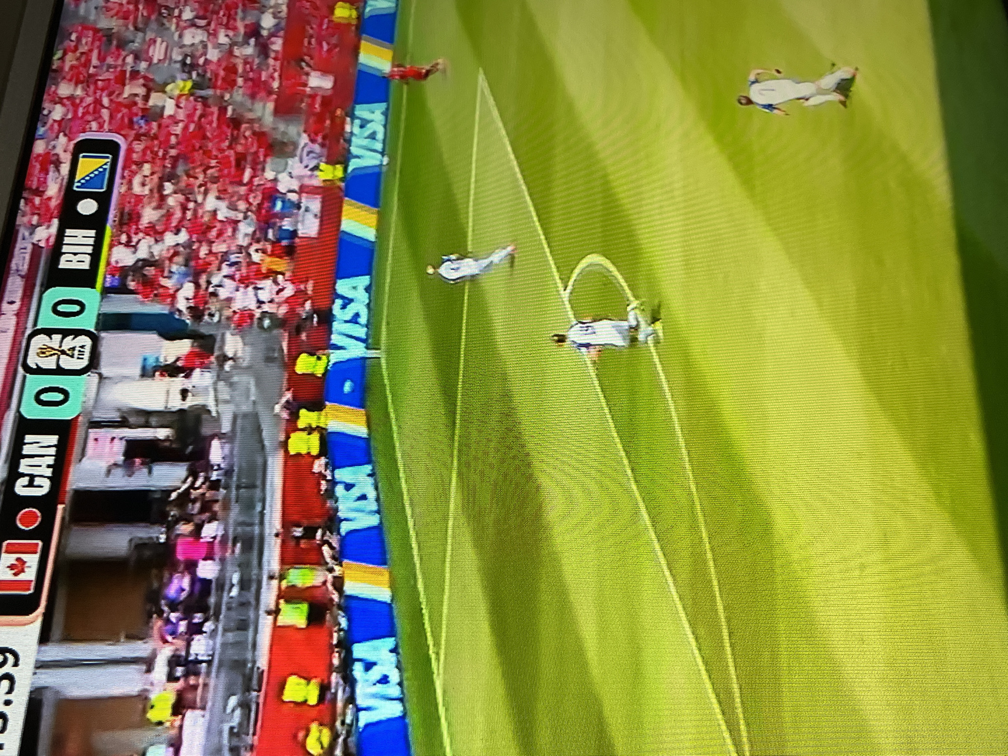 This is a photograph of a television screen displaying a soccer match. The scoreboard at the top indicates the match is between Canada (CAN) and Bosnia and Herzegovina (BIH), with the score currently 0-0. The players on the field are wearing white and blue uniforms. The field is green with white lines, and the background shows a stadium filled with spectators wearing red and white. Several "VISA" logos are visible along the sideline of the field. The overall lighting suggests an indoor or evening game. The image does not provide any information about the specific city or location of the match.