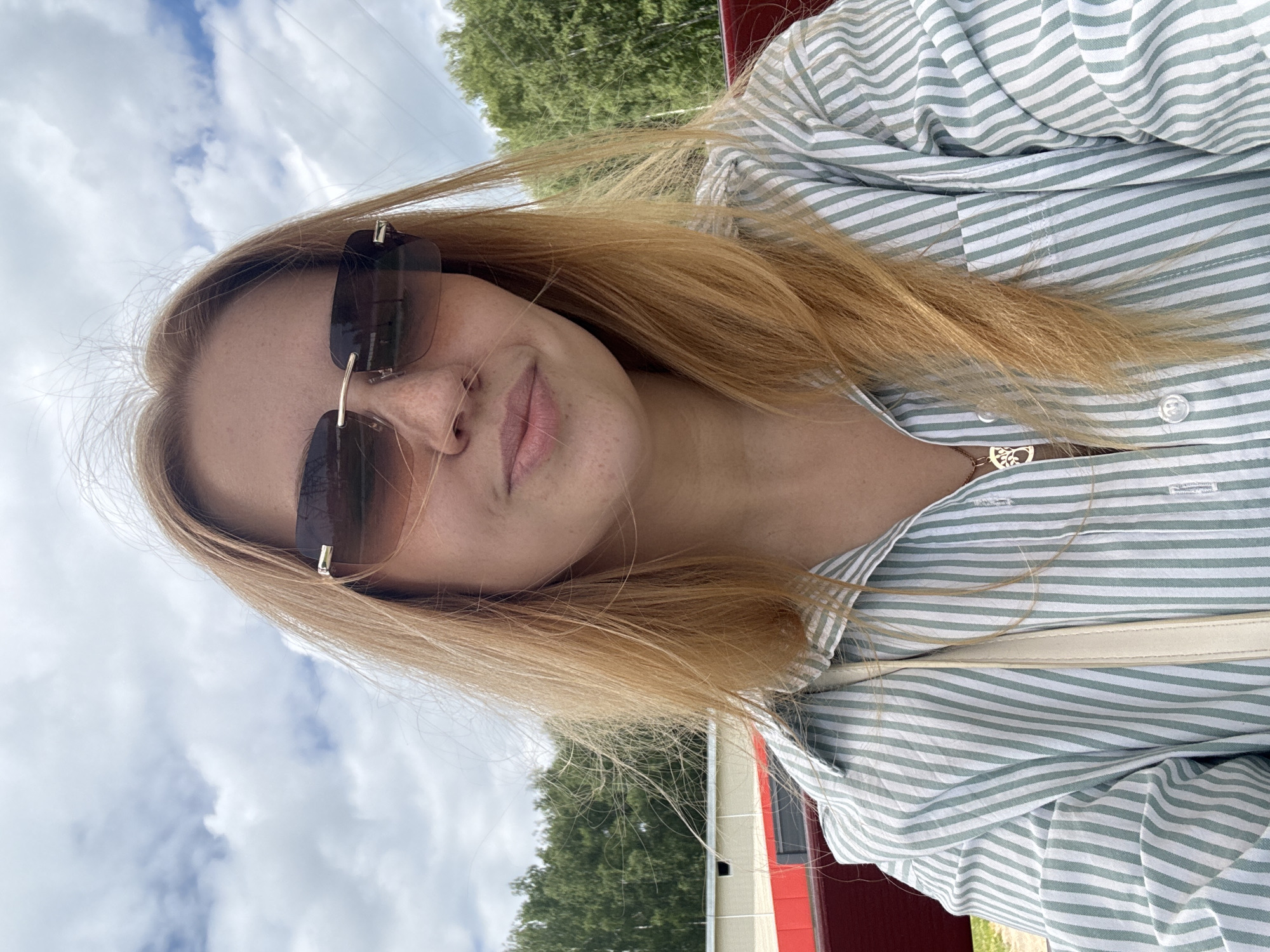 The media file presents a rotated close-up selfie of a young woman, captured outdoors. The image is rotated approximately 90 degrees counter-clockwise, showing the subject's head to the left and her body extending downwards.
The primary subject is a young woman with long, straight blonde or light brown hair that partially covers her shoulder and chest. She is wearing dark, rectangular sunglasses that obscure her eyes. Her face is turned slightly towards the camera, revealing freckles across her nose and cheeks. Her lips are closed in a neutral or slightly pleasant expression.
She is dressed in a button-up shirt featuring light green and white stripes. A delicate gold-toned necklace with a detailed, tree-shaped pendant is visible around her neck, resting on her collarbone. A light-colored strap, possibly from a shoulder bag or purse, can be seen draped across her front.
The background indicates a bright, cloudy daytime setting. The sky, filled with white and grey clouds, occupies the upper part of the frame. Below the sky, green foliage from trees is visible, suggesting a natural, outdoor environment. A portion of a red building or structure is also discernible on the right side of the image. Faint power lines can be seen in the sky against the clouds. There is no visible text in the image. The location context provided is Yaroslavl, Russia.