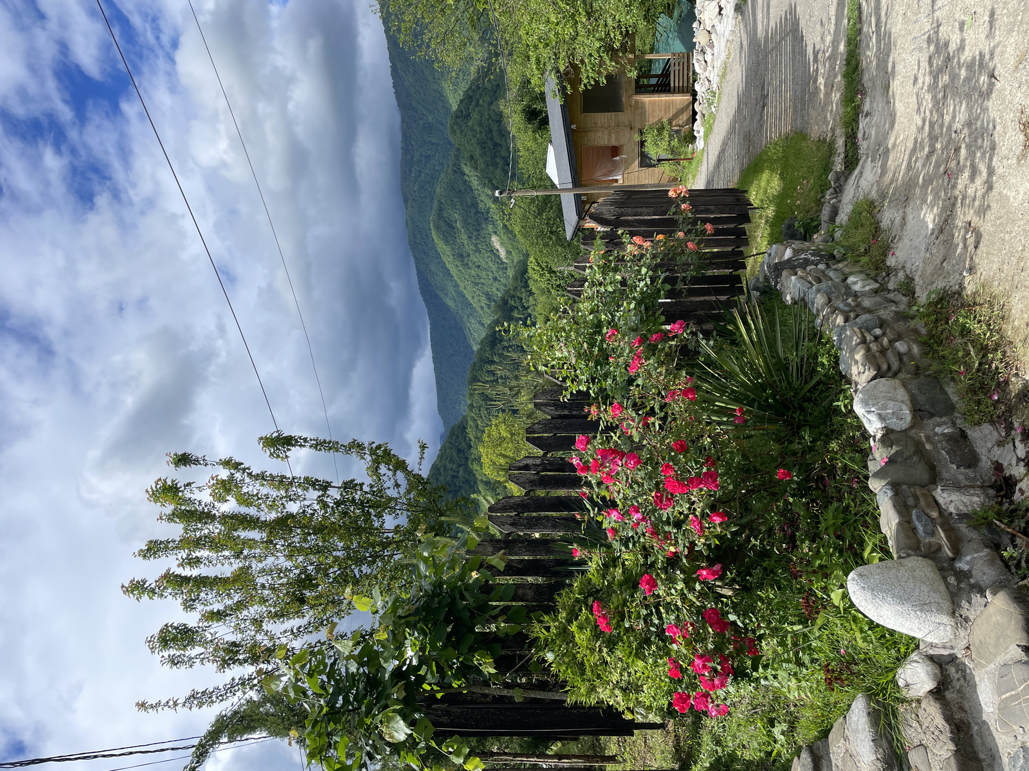 The image shows a scene in Solokh-Aul, Russia. In the foreground, a wooden fence is partially covered with lush greenery and vibrant red flowers. Adjacent to the fence is a stone wall constructed with various sized stones. Beyond the fence, a paved pathway leads towards a simple wooden building. The background features rolling green mountains under a sky filled with dramatic clouds; patches of blue sky are visible between the clouds. Power lines run across the upper left corner of the image.