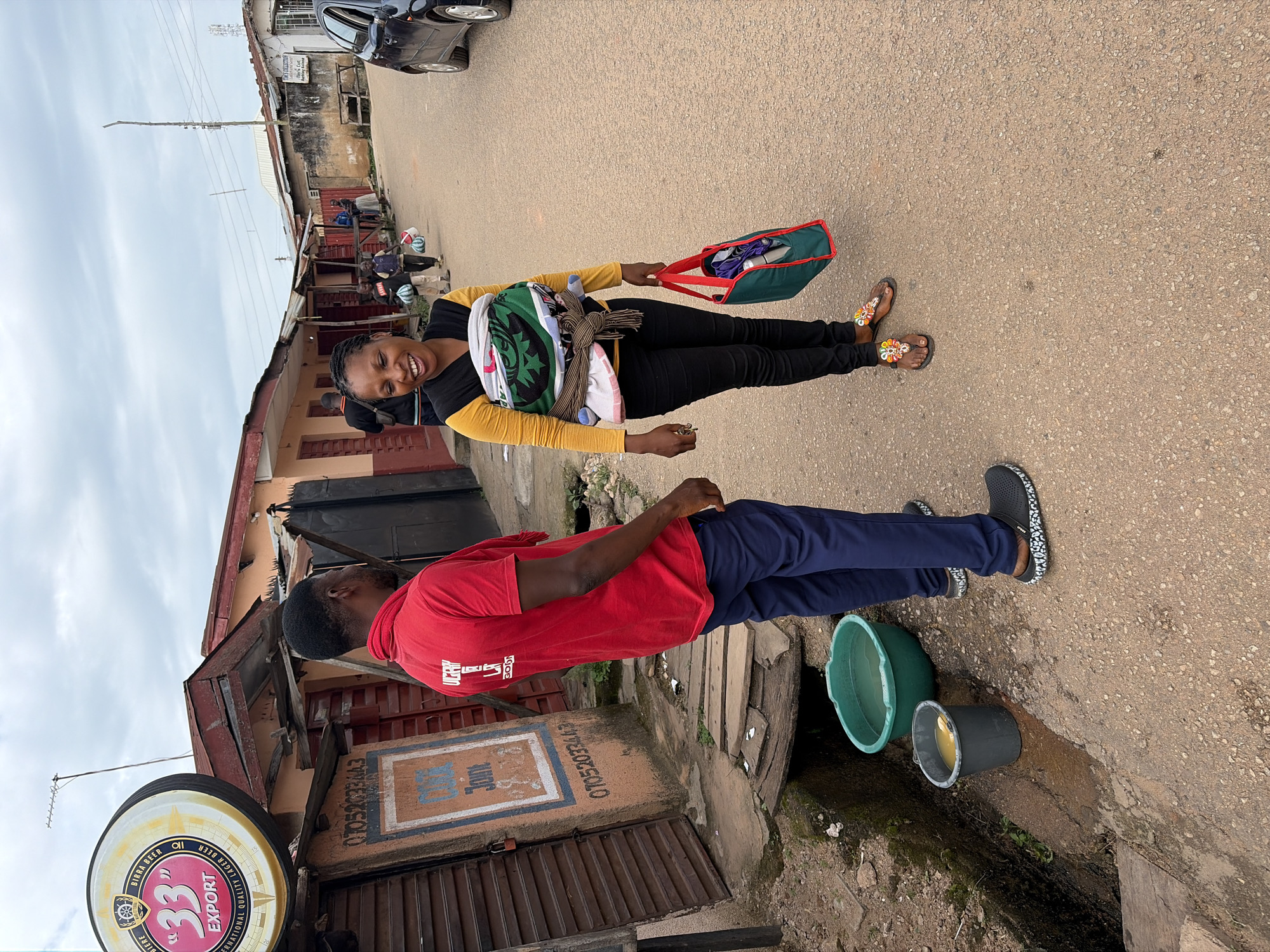 In Jos, Nigeria, a street scene unfolds under an overcast sky. A man in a red t-shirt and navy blue pants stands facing a woman who is smiling broadly and carrying a baby wrapped in a colorful cloth on her back. The woman is wearing a yellow long-sleeved shirt and black pants, with beaded sandals on her feet. She holds a green and red tote bag in her right hand.
The background features a row of buildings with corrugated iron roofs, some appearing weathered. A large circular sign for "33 EXPORT" beer is prominently displayed on the left, with a phone number "07052032443" visible on a nearby wall. Several people are walking in the background, and a dark-colored car is parked on the right side of the street. In the foreground, a green plastic basin and a gray bucket, both containing liquid, are placed near a drainage ditch. The ground is paved with asphalt and gravel.