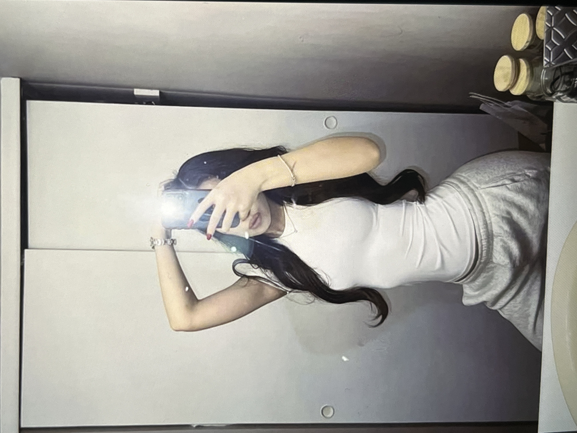 A person is taking a mirror selfie in what appears to be a closet or dressing area. The person is wearing a white tank top and light gray sweatpants. They have long, dark hair and are holding a phone in their right hand, which is raised to capture the reflection. The person is also wearing a bracelet on their left wrist and has red nail polish. In the background, there are two large sliding doors, likely closet doors, and a glimpse of a countertop with some jars and a patterned object. The lighting in the room is somewhat dim, with a lens flare visible from the phone's camera. The overall environment suggests a private, domestic setting.