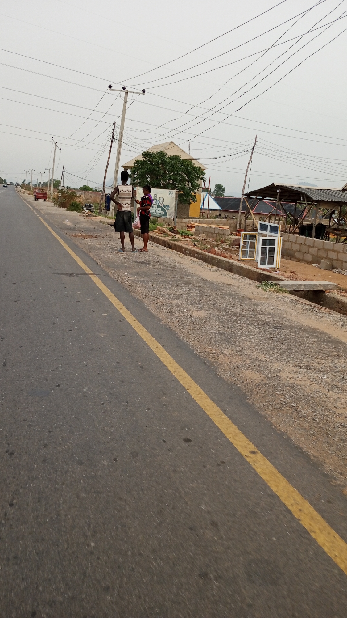 The image depicts a street scene in Adankolo, Nigeria. A yellow line runs along the left side of a two-lane asphalt road. On the right shoulder of the road, two individuals are standing; one is wearing a striped shirt and dark shorts, while the other is wearing a patterned purple shirt and dark shorts, holding a bag. Next to them is a sign, and further down the shoulder are building materials, including bricks, and framed glass windows. A partially constructed wooden structure with a roof is also visible. Overhead, numerous power lines crisscross the sky, supported by utility poles. A red vehicle is parked on the opposite side of the road in the distance. The weather appears overcast.