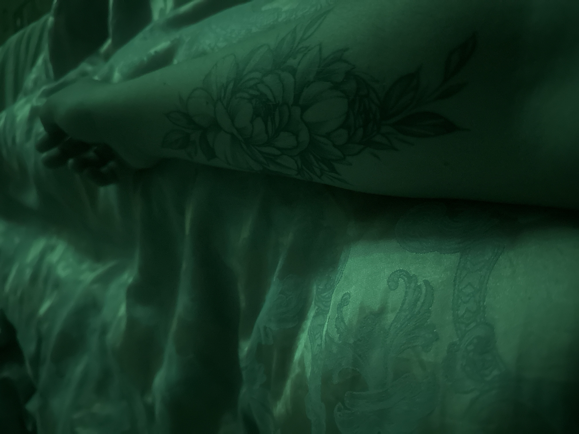 The image shows a close-up of a person's arm resting on a green, textured fabric, possibly a bedspread or blanket. The fabric has an embossed or patterned design visible in the light. A tattoo of a floral design, featuring a large bloom and leaves, is visible on the person's forearm. The lighting is dim and casts a green hue over the scene, obscuring some details. There are no visible indications of the location being in Gavrilov Posad, Russia, or any urban environment.
