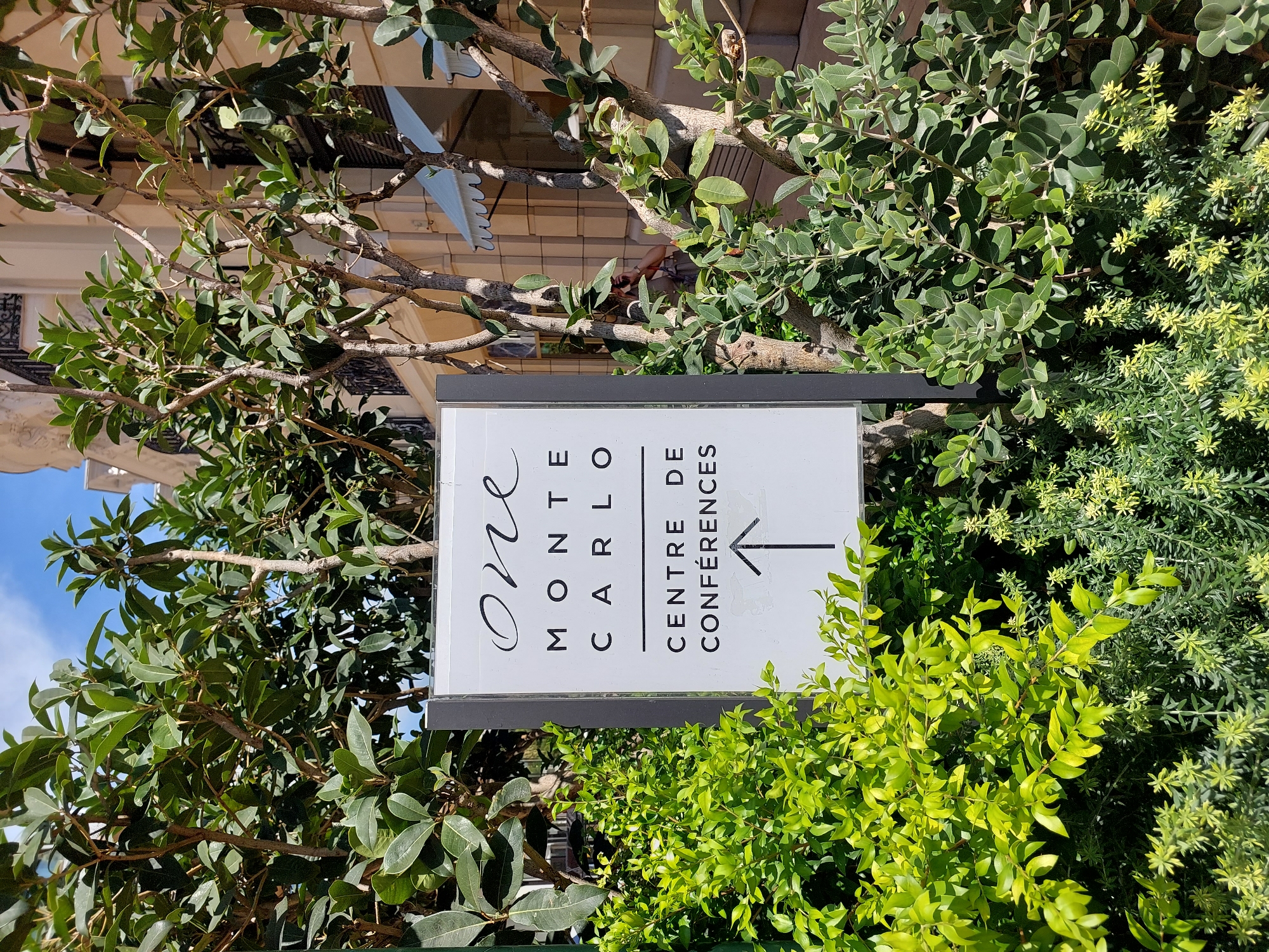 The image shows a sign in front of the Grimaldi Forum in Monaco, which is a convention center in Monaco. The sign reads "ONE MONTE CARLO CENTRE DE CONFERENCES" with an arrow pointing upwards, indicating the direction to the conference center. The sign is surrounded by lush greenery and trees. In the background, the building of the Grimaldi Forum is visible, with its distinctive architecture. The sky is blue with some clouds, suggesting a clear day.