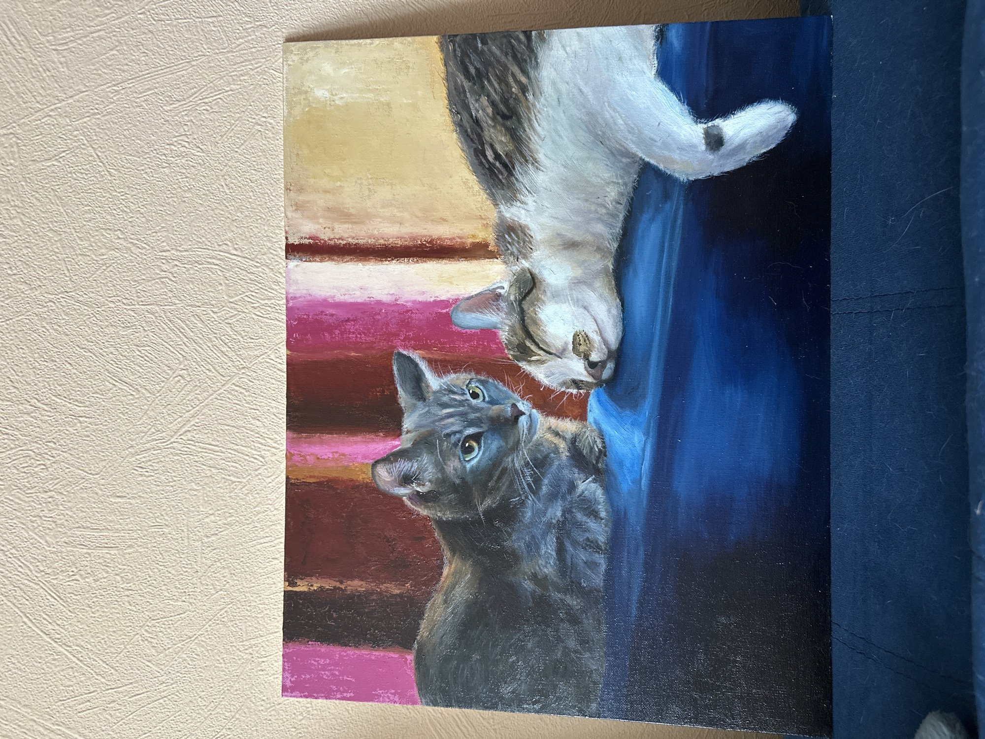 The image shows a painting of two cats. One cat is grey and looking up at the other, which is white and tabby, hanging upside down. The background of the painting is comprised of horizontal stripes in shades of pink, red, and yellow on one side and a vertical blue panel on the other. The painting appears to be hanging on a beige textured wall, next to a dark blue fabric. The image does not contain any information about the city of Krasnoyarsk, Russia.
