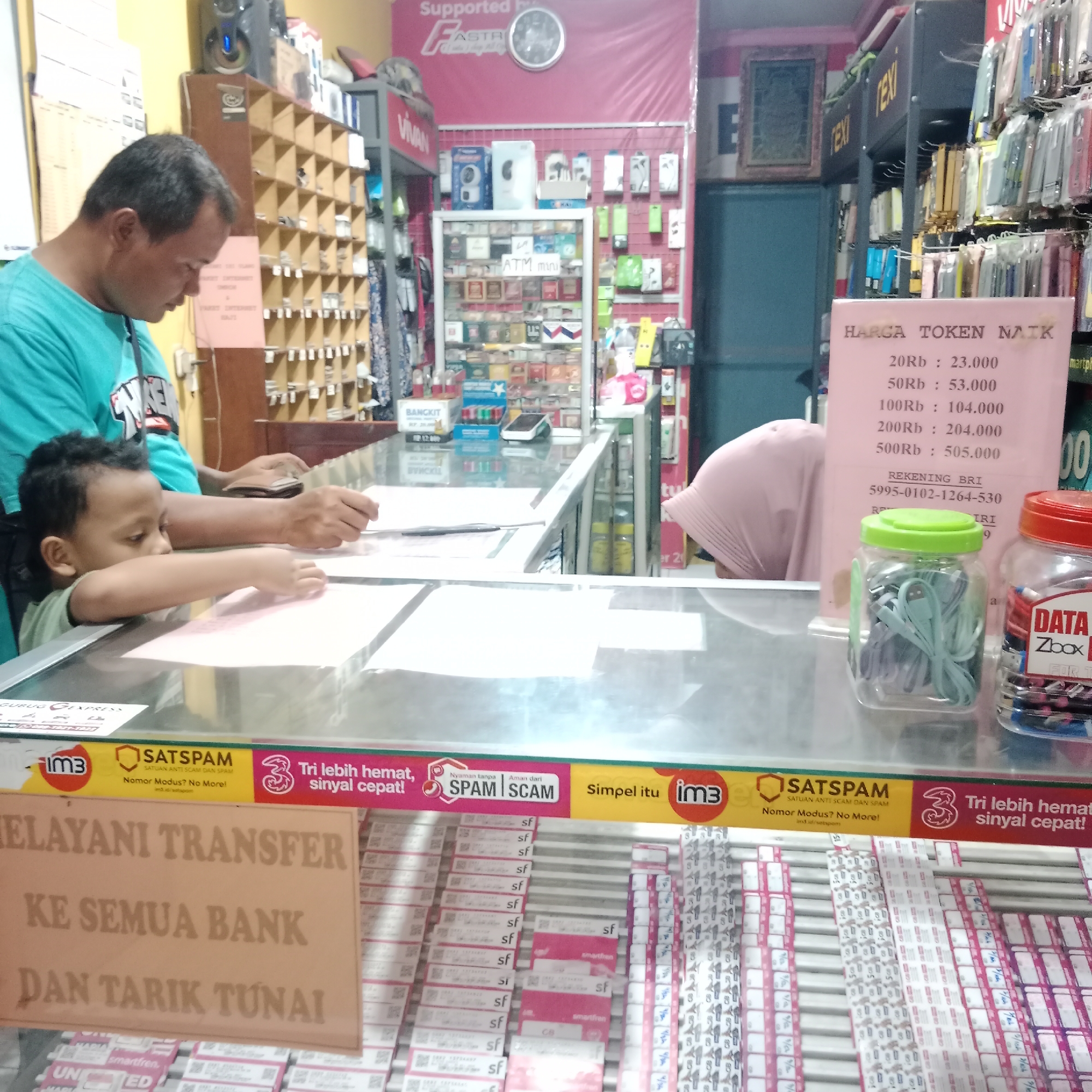 The image shows a storefront in Tegowanu Kulon, Indonesia, likely a mobile phone and accessories shop. In the foreground, a man in a teal t-shirt is leaning over a counter, seemingly engaged in a transaction with a child. The child, wearing a light green shirt, is also leaning on the counter, reaching towards papers. The counter itself is glass-topped, revealing various items below, including stacks of what appear to be SIM cards or phone credit vouchers.
Behind the counter, shelves are stocked with a variety of products, including phone cases, electronic accessories, and what seems to be a display of various brands of mobile phone starter packs or data packages. A sign in pink hangs in the mid-ground with text that appears to be pricing information for "TOKEN NAIK" (token increase) with various denominations and prices listed. Another sign in the foreground, also in a light brown color, states "MELAYANI TRANSFER KE SEMUA BANK DAN TARIK TUNAI" (Serving transfers to all banks and cash withdrawals).
The environment suggests a busy retail setting with items densely displayed. Branding elements are visible, including "im3" and "SATSAPAM" (anti-scam and spam unit) logos. The overall impression is of a typical Indonesian small business operating in a local community.